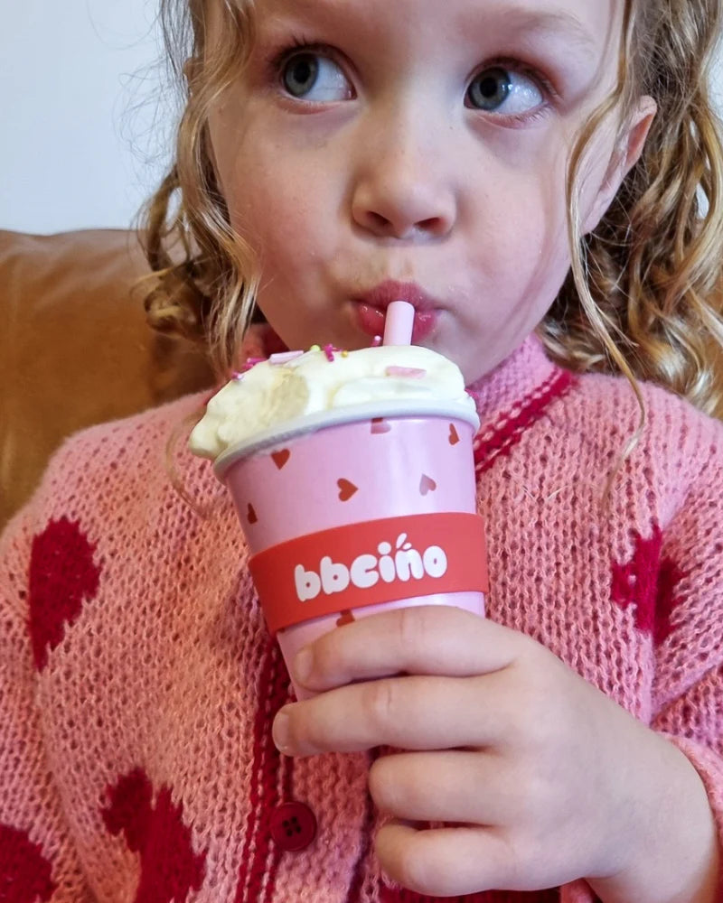 A young child with curly hair sips thoughtfully from a 120ml BBCINO Reusable Babycino Cup Sweetheart, which is pink and decorated with hearts. The child uses a silicone straw and wears a pink sweater featuring darker pink heart patterns, opting for an eco-friendly alternative to regular babycino cups.