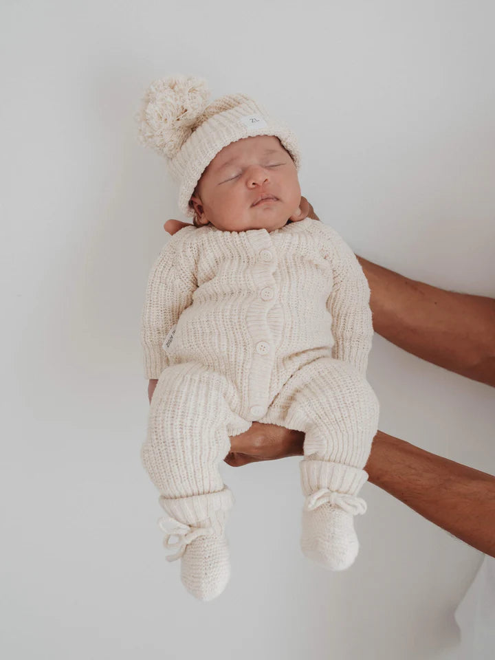 A baby dressed in the ZIGGY LOU Classic Knit Romper Honey and matching hat with a pom-pom is being held up by an adult's hands against a plain background. The winter outfit keeps the little one snug and cozy as they sleep peacefully, their closed eyes and serene expression adding to the charm.