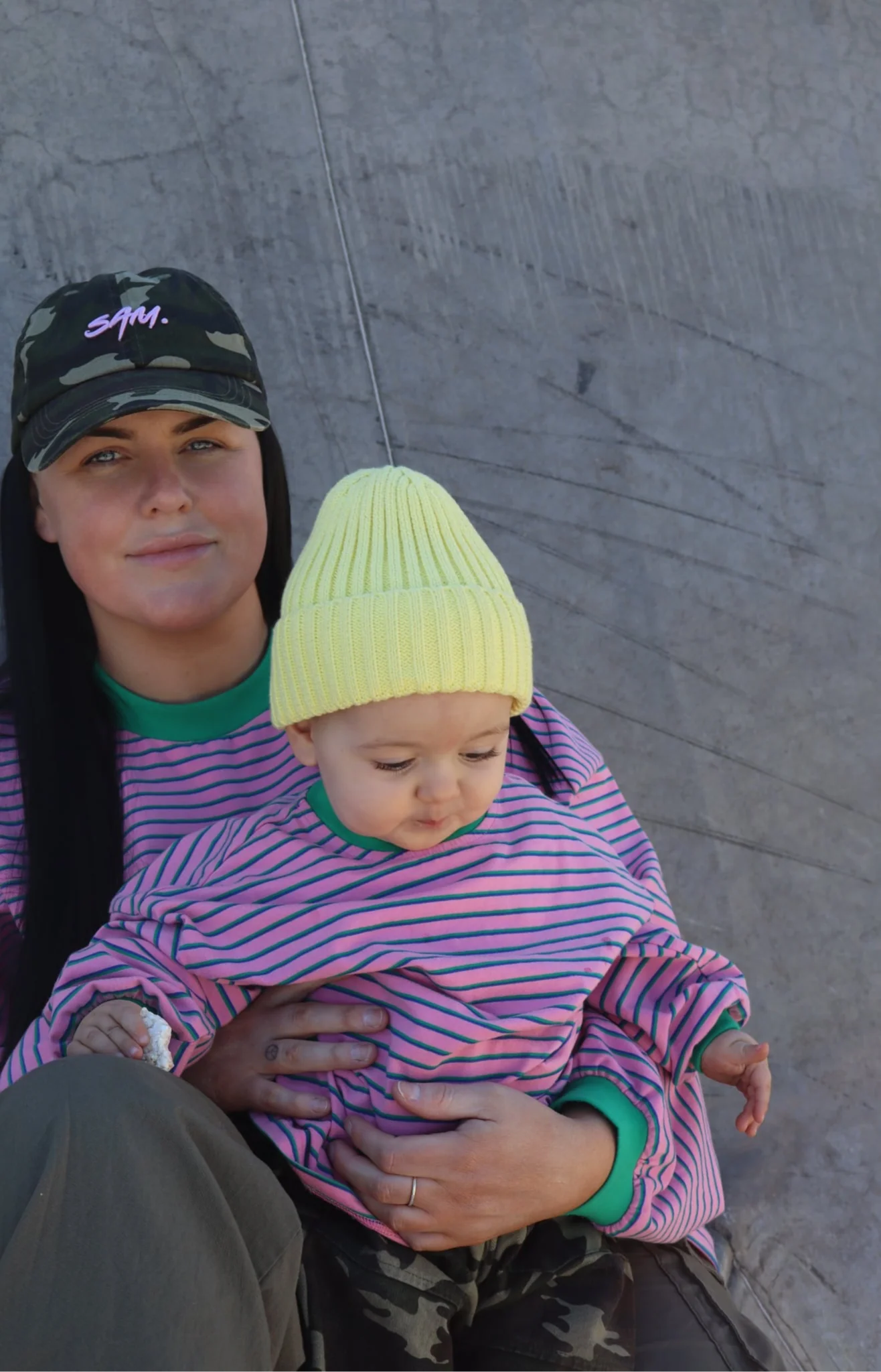 A woman in a camo hat and striped pink and purple shirt holds a baby wearing the SAM THE LABEL Kids Watermelon Sugar LS Tee and a yellow knit hat, as they sit against a gray concrete wall.
