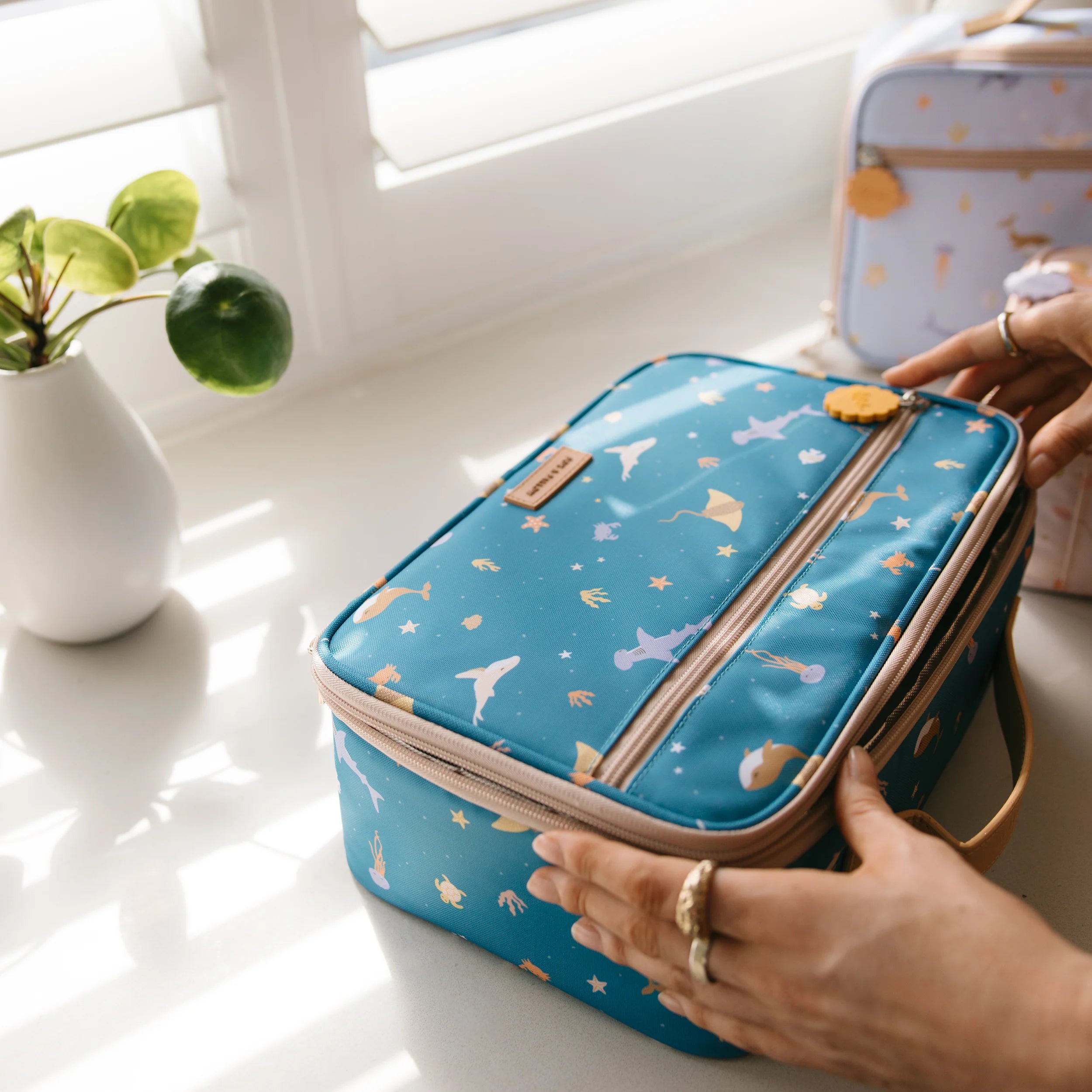 Two hands are holding the FOX & FALLOW Ocean Creatures Marine Lunch Bag, which features a whimsical print of animals and umbrellas on a blue background. Made from waterproof fabric, this insulated lunch bag rests on a white countertop with sunlight streaming through nearby window blinds. A small potted plant sits to the left, and another lunch bag is visible in the background.