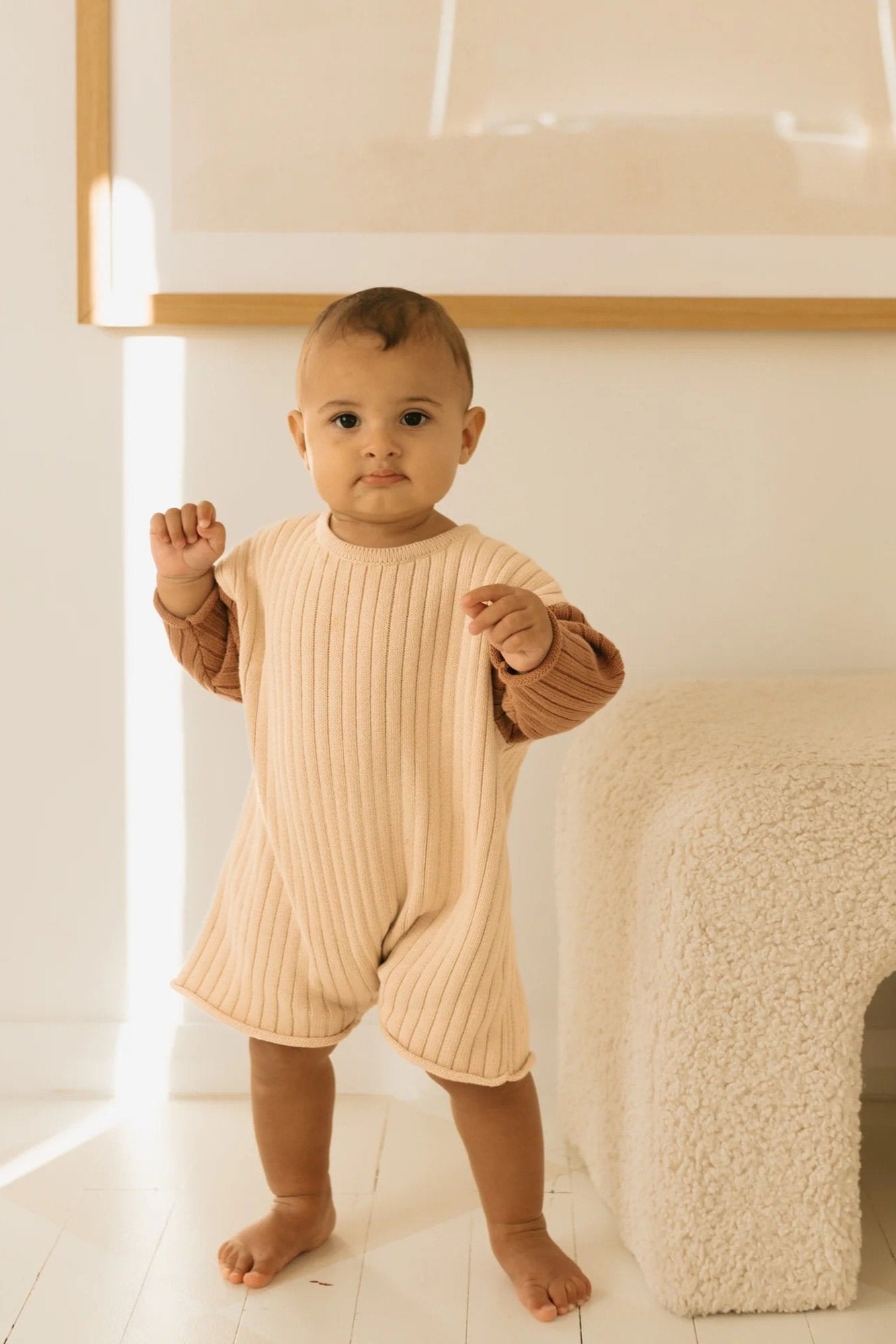 A baby stands on a light wooden floor in a bright room, dressed in the Golden Knit Romper Caramel Sundae from GOLDEN CHILDREN. The baby is barefoot, and a soft, light beige bench is positioned to the right. The room features minimalist decor with framed artwork on the wall.