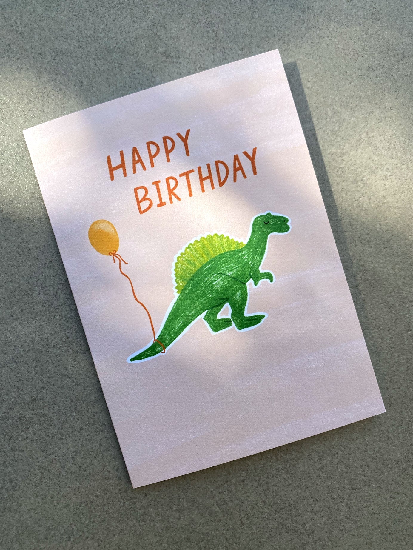 The "Birthday Greeting Card 'Dinosaur Balloon'" from LAUREN SISSONS STUDIO features a whimsical dinosaur illustration accompanied by an orange balloon on the front. Vibrant orange letters at the top of the card spell out "HAPPY BIRTHDAY." This delightful card is printed on recycled paper and is set against a gray surface.