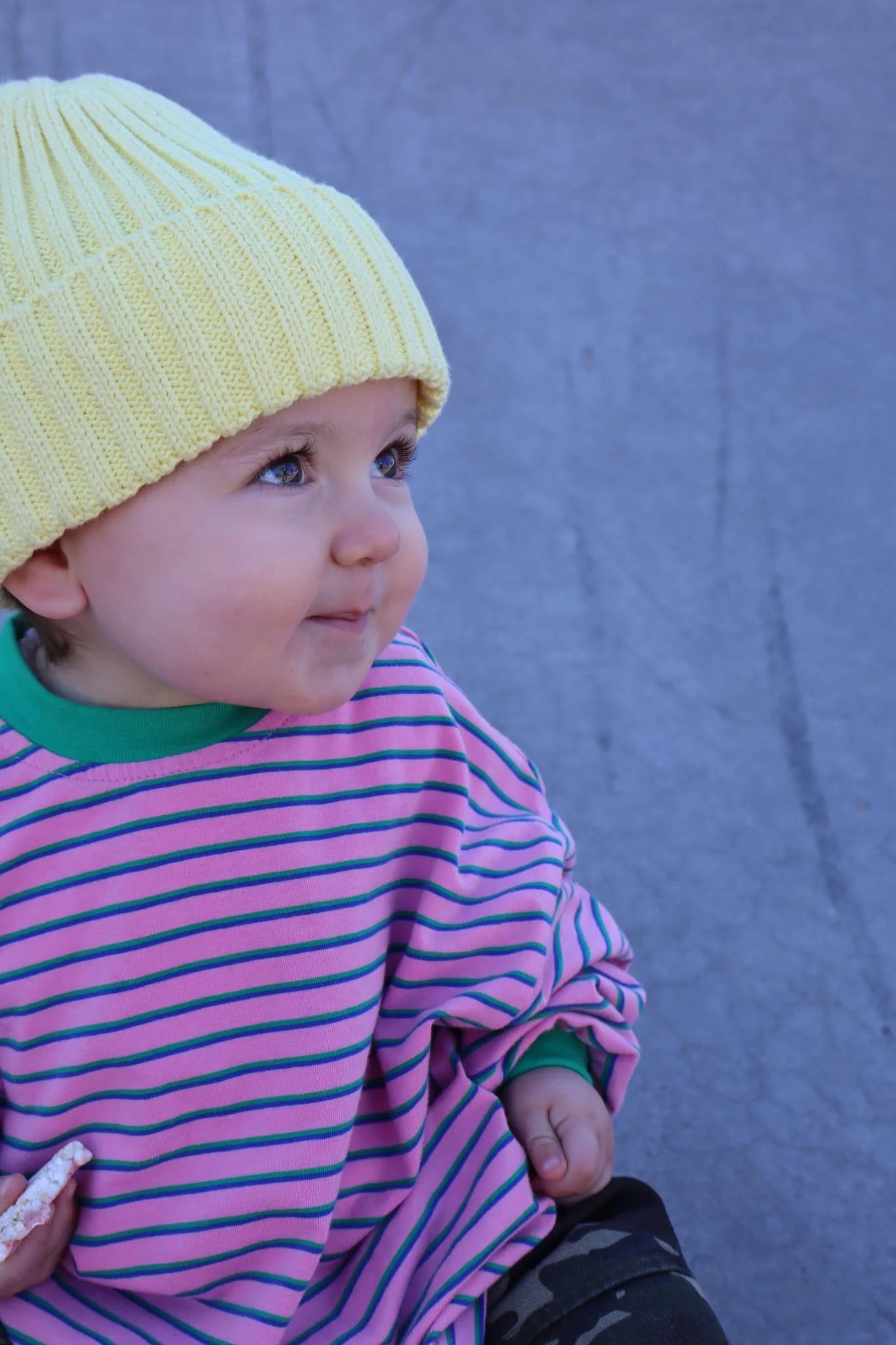 A baby wearing a yellow knit hat and the Sam the Label - Kids Watermelon Sugar LS Tee by SAM THE LABEL, featuring pink-and-blue stripes, looks up with a slight smile while sitting outdoors on a gray surface.