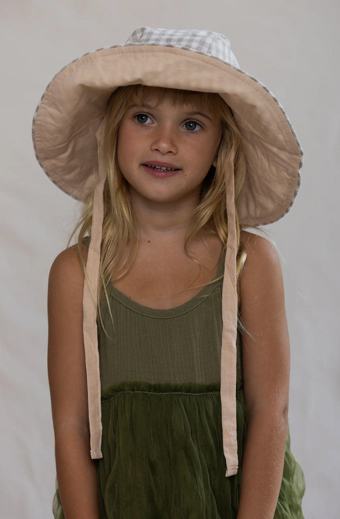 A young girl with long blonde hair is wearing a brimmed hat with a checkered pattern on the inside and a solid color on the outside. She is dressed in the Bangalow Tutu Dress Eucalyptus by VALENCIA BYRON BAY and looking slightly to the side with a neutral expression. The background is plain.