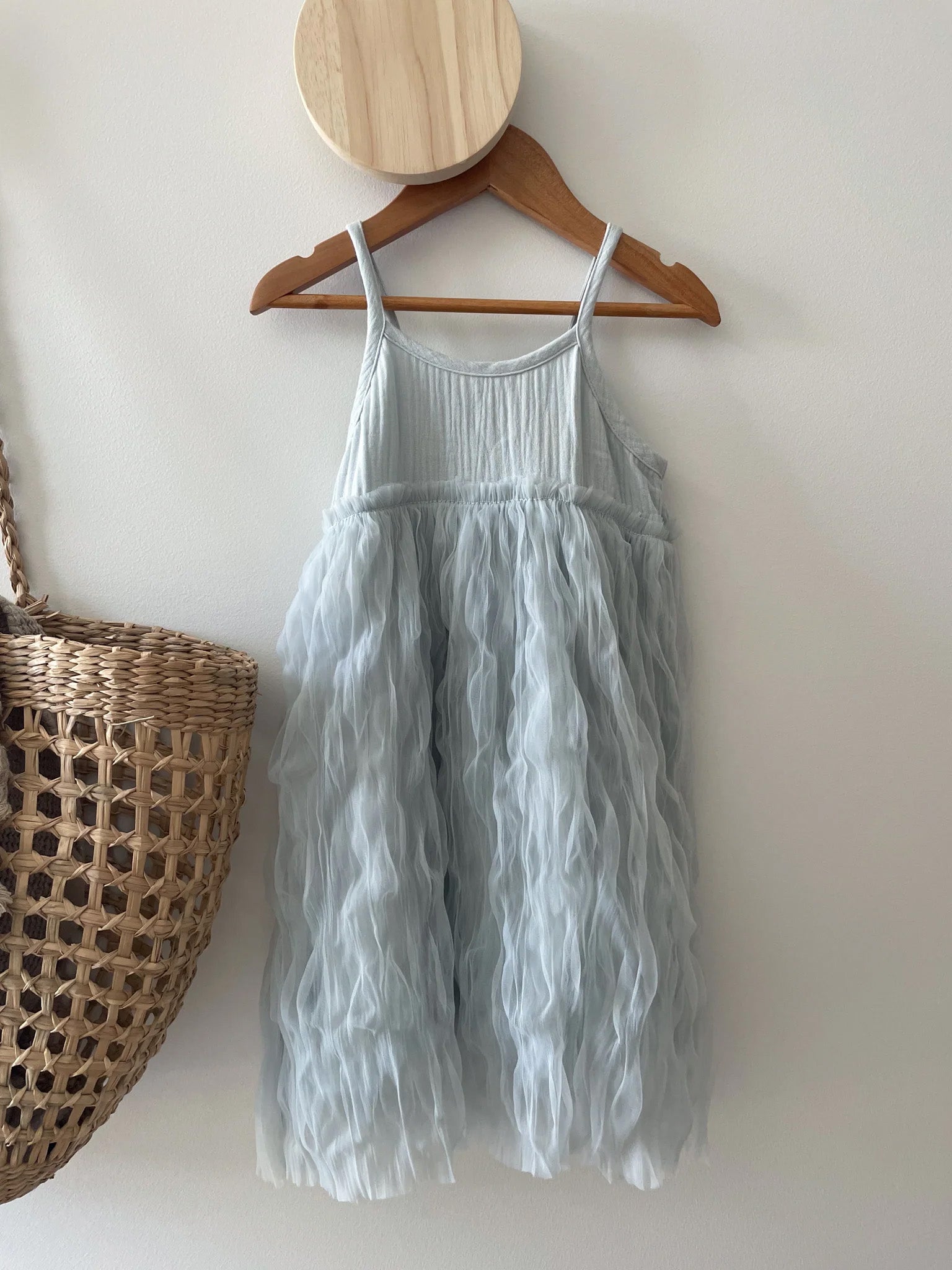 A charming dusty blue, sleeveless Bangalow Tutu Dress Elsa from VALENCIA BYRON BAY, featuring a ruffled tulle skirt reminiscent of Frozen’s Elsa, hangs gracefully on a wooden round hanger against a white wall. Nearby, an empty woven basket also adorns the wall. Light shadows dance across the surface.