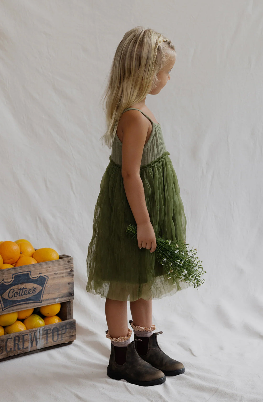 A young girl with long blonde hair stands in profile, adorned in a green Bangalow Tutu Dress Eucalyptus by VALENCIA BYRON BAY and black boots. In her right hand, she holds a small bouquet of greenery. Behind her, a wooden crate filled with oranges and lemons is partially visible against a plain white background.