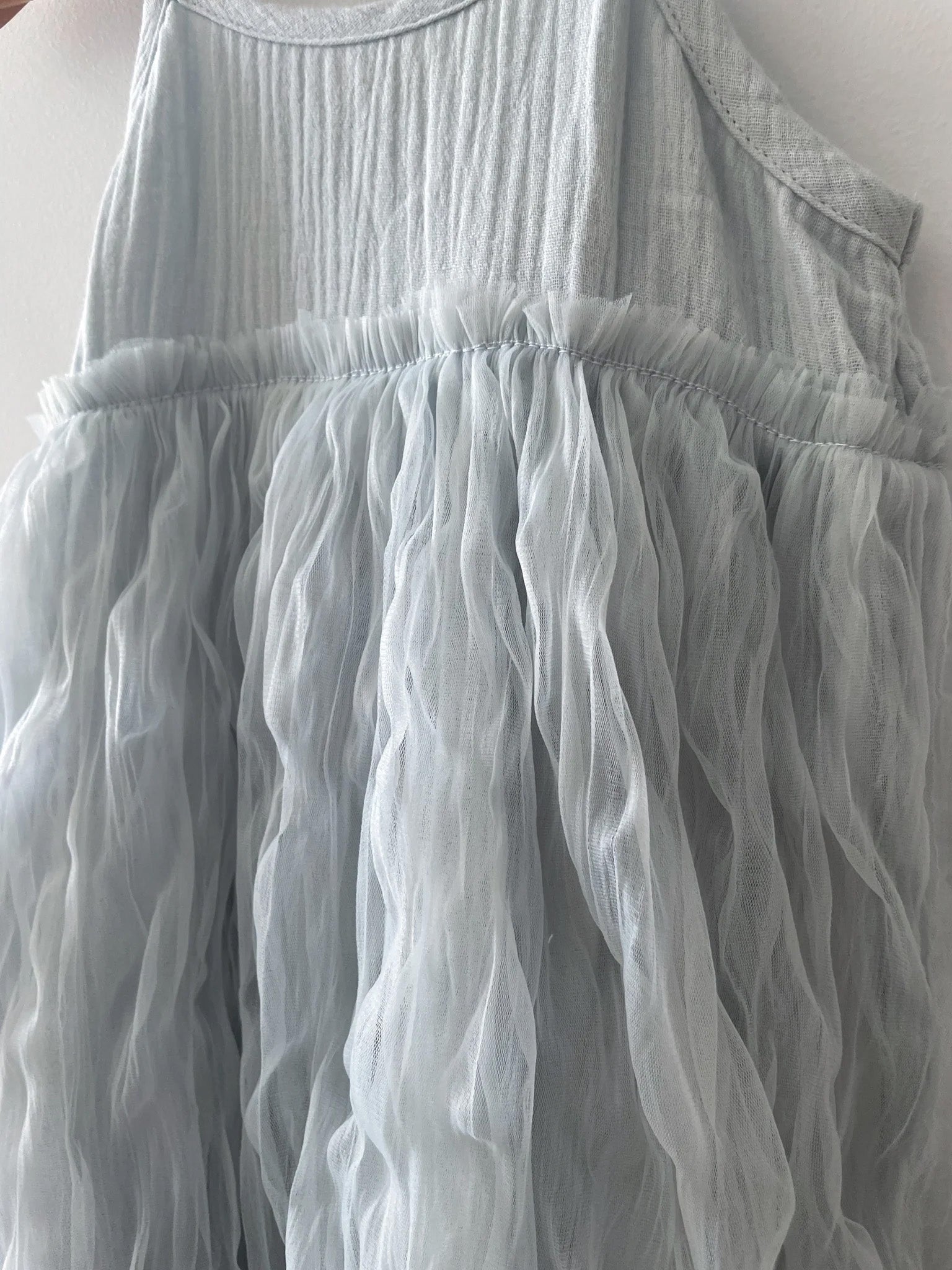 A close-up view of the Bangalow Tutu Dress Elsa by VALENCIA BYRON BAY, showcasing its pale, dusty blue shade. The sleeveless dress features a gathered and ruched tulle skirt with a crinkled texture, creating a wavy, airy appearance reminiscent of Frozen character Elsa's ethereal style.