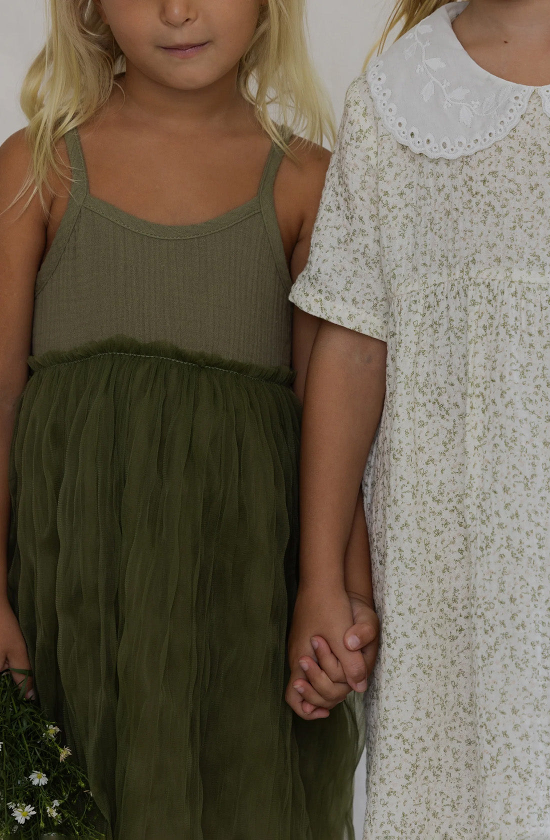 Two young children holding hands. The child on the left is wearing a stunning Bangalow Tutu Dress Eucalyptus by VALENCIA BYRON BAY and holds a small bouquet of flowers, while the one on the right is in a white party dress with a floral pattern and a Peter Pan collar.