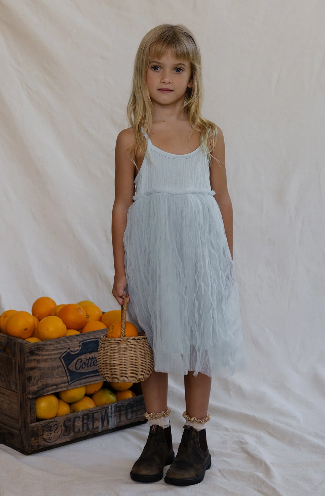 A young girl with blond hair is wearing a white sleeveless dress that evokes the look of the Bangalow Tutu Dress Elsa by VALENCIA BYRON BAY, paired with dark boots. She is holding a small wicker basket and standing next to a wooden crate filled with oranges. The background features a simple, white, draped cloth that channels the elegance of Frozen character Elsa's style.