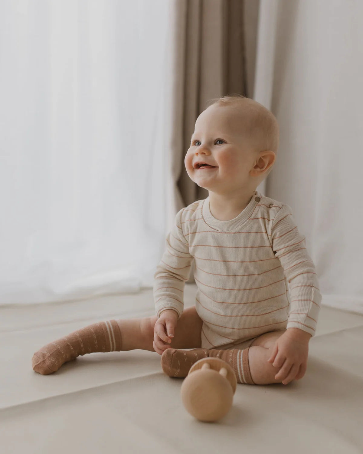 A happy baby with light hair sits on the floor in a cozy, well-lit room. The baby is wearing the SUSUKOSHI Classic Bodysuit Coco Stripe, ethically made in China, paired with brown knee-high socks. The baby is holding a wooden toy, smiling and looking upward. White and beige curtains are visible in the background, enhancing the serene setting.