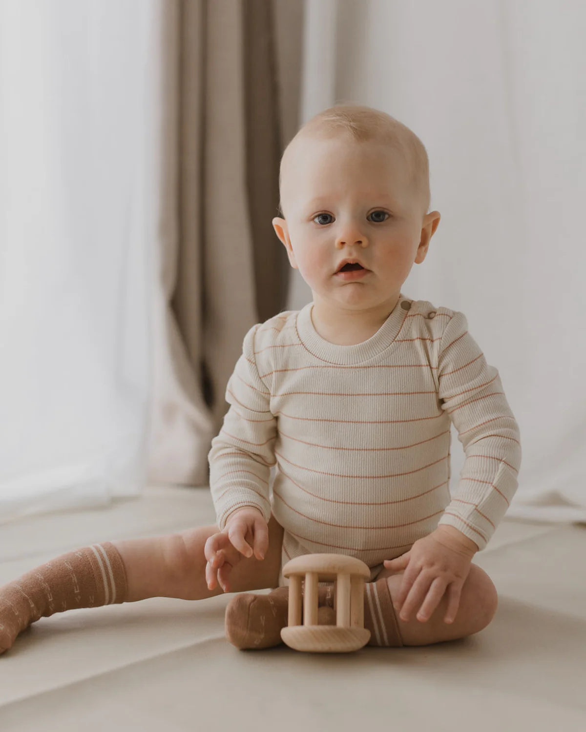 A baby with short blond hair sits on the floor wearing the SUSUKOSHI Classic Bodysuit Coco Stripe, a light-colored, long-sleeved onesie with thin horizontal stripes and matching socks, all crafted from organic cotton. The baby holds a wooden toy and is surrounded by soft, neutral-toned curtains in the background.