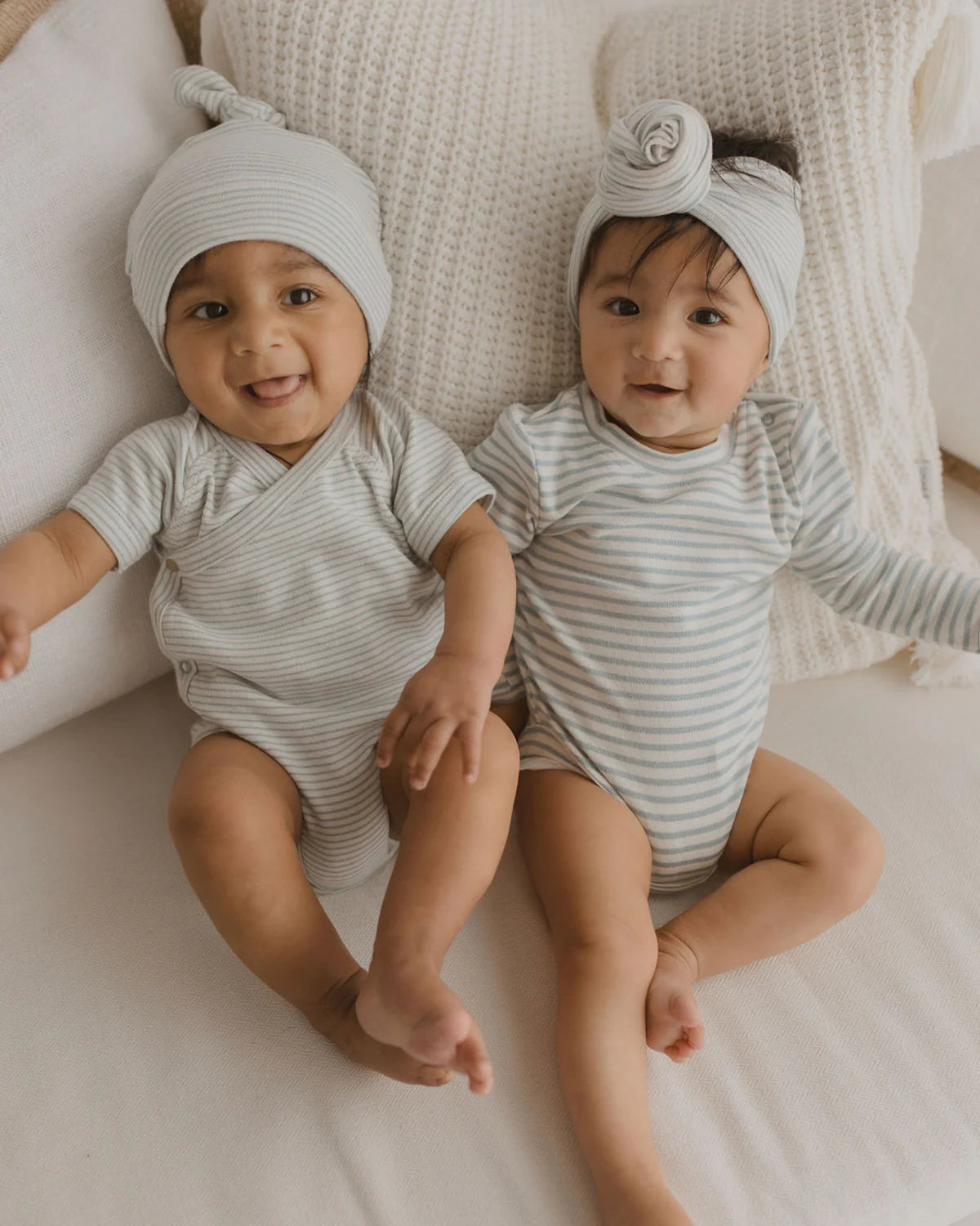 Two infants, wearing matching SUSUKOSHI Classic Bodysuit Seaside in light gray stripes, sit close together on a cream-colored couch. Both are adorned with light gray caps; one featuring a playful knot and the other embellished with a delicate fabric rose. They are smiling, appearing happy and relaxed, with a textured white pillow behind them.