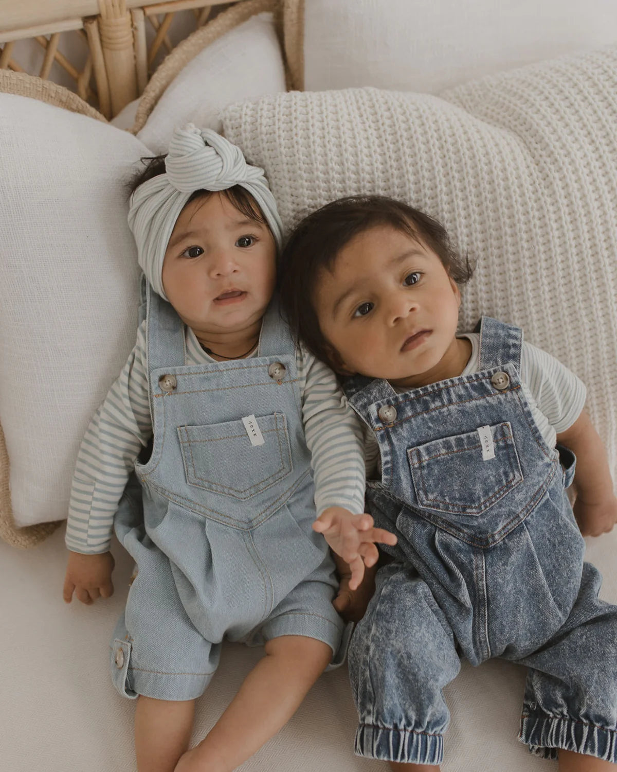 Two babies sit on a couch against white cushions. One baby wears a light denim overall with a striped shirt and a knotted headband, while the other is dressed in darker denim overalls with a striped shirt. Both have dark hair and look towards the camera, showcasing their adorable Classic Bodysuit Seaside by SUSUKOSHI, ethically made for maximum baby comfort.