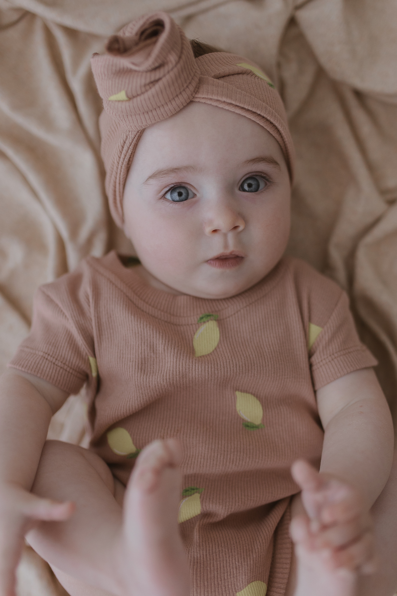 A baby with blue eyes lies on a beige blanket, dressed in the SUSUKOSHI Classic Bodysuit Lemons. This peach-colored outfit adorned with lemon prints comes with a matching headband and is made from organic cotton, ensuring comfort as the baby's hands and feet are slightly raised.