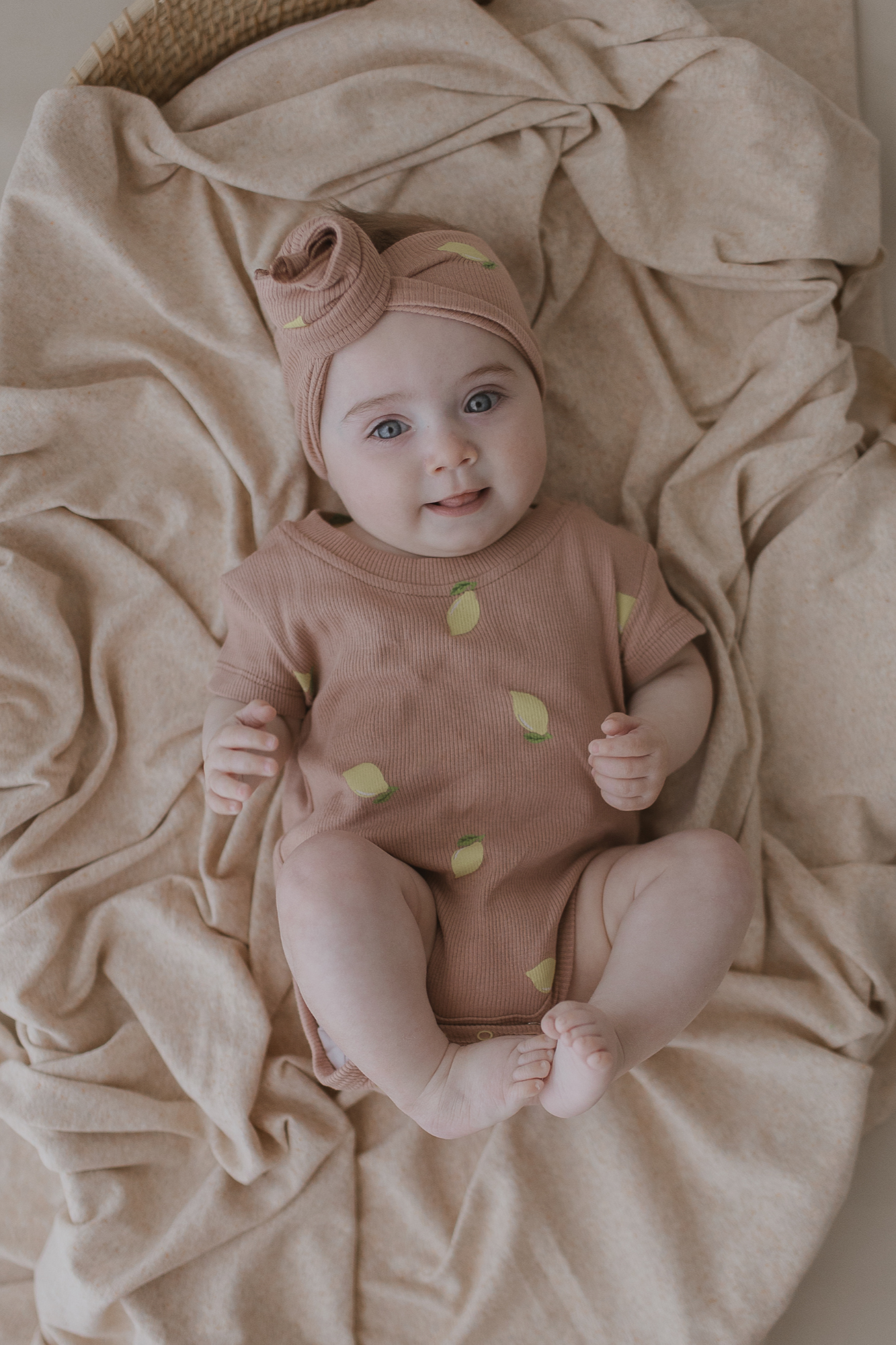 A baby is wearing a matching headband and the Classic Bodysuit Lemons from SUSUKOSHI, lying on a soft beige blanket, gazing upward with a calm expression.