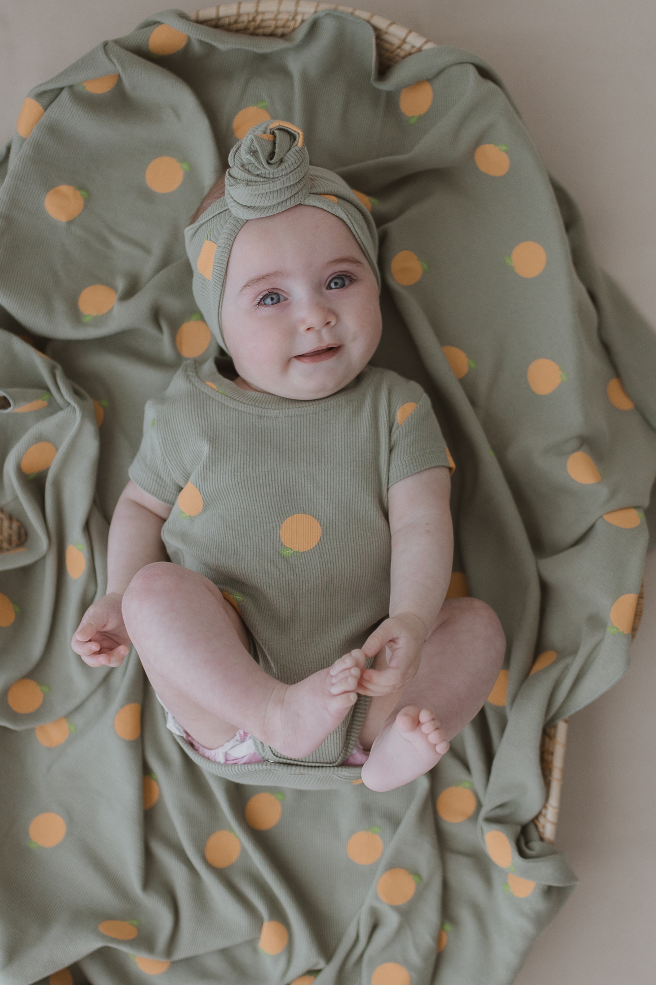 A smiling baby in the SUSUKOSHI Classic Bodysuit Oranges, made from ethically sourced organic cotton, lies on matching fabric. The green outfit with yellow polka dots includes a coordinating headband, and the baby rests adorably in a wicker basket, highlighting charming and comfortable baby attire.