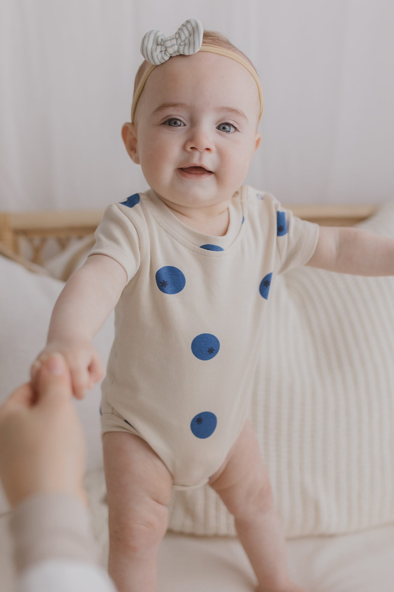 A smiling baby wearing the Classic Bodysuit Blueberries by SUSUKOSHI, crafted from ethically sourced materials, is supported to stand by unseen adult hands. The cozy, neutral-toned setting with soft pillows enhances the charm of this delightful moment.