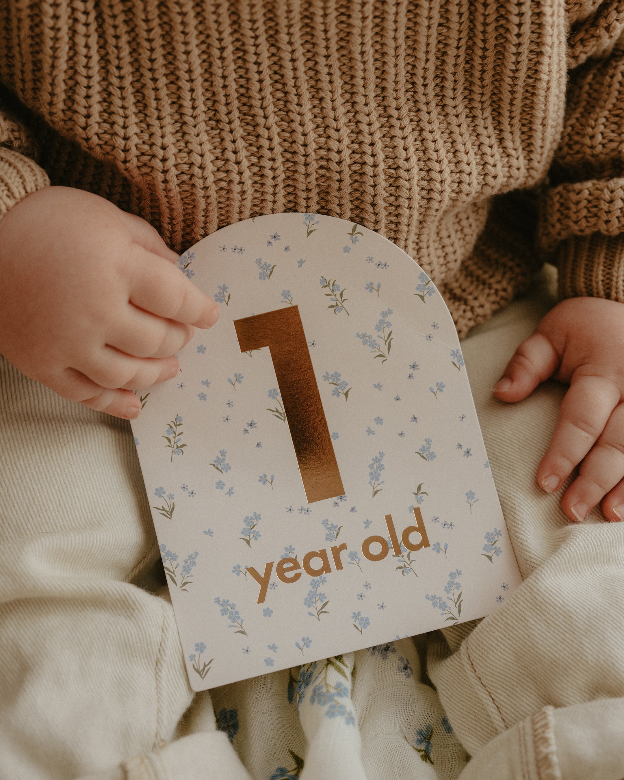 A baby in a knit sweater and light-colored pants holds a FOX & FALLOW Baby Milestone Cards Broderie sign with the text "1 year old" in gold lettering. The sign has a floral pattern with small blue flowers and green leaves. The baby's face is not visible.