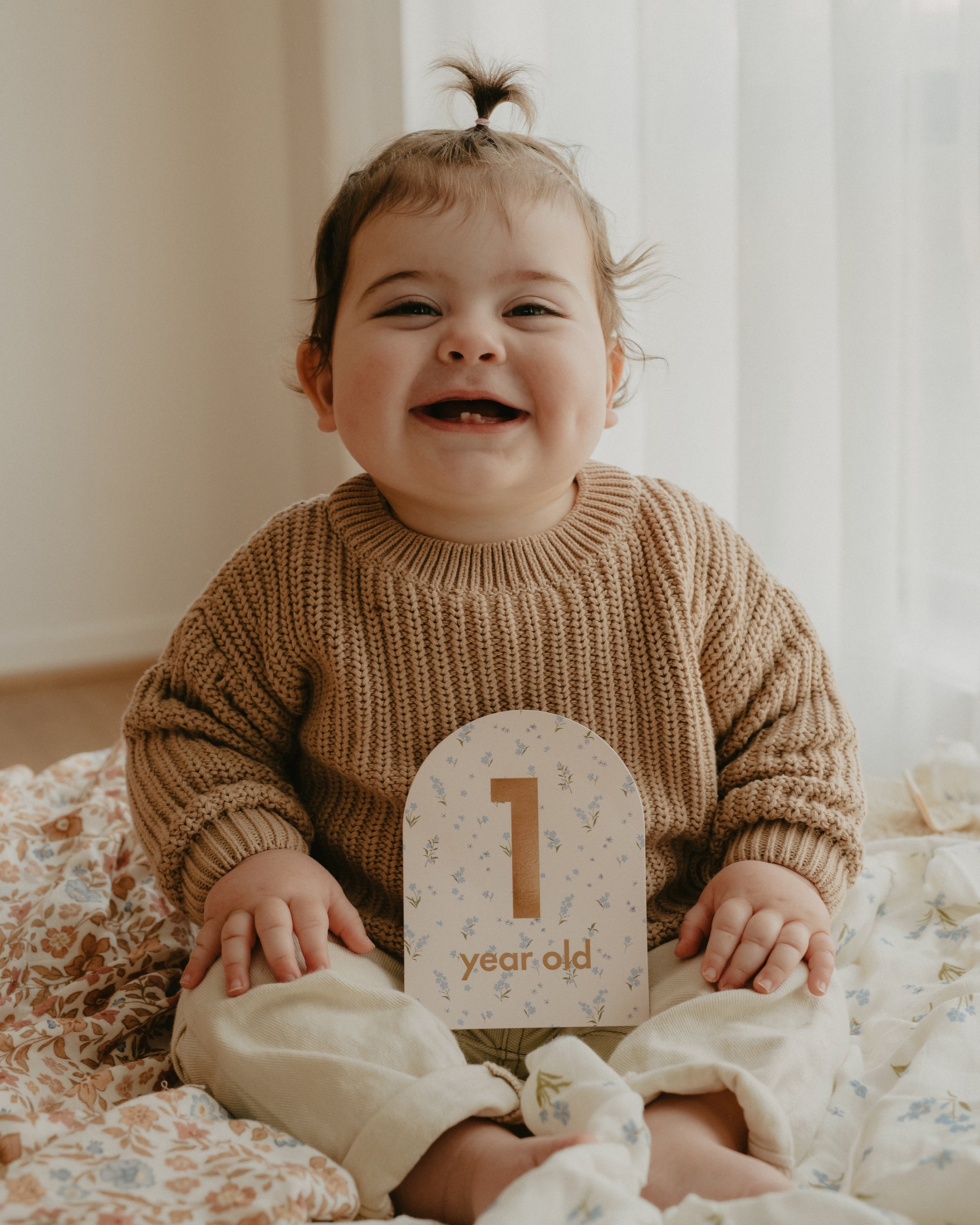 A happy baby with dark hair tied in a small ponytail sits on a decorative quilt. The baby is wearing a light brown sweater and cream-colored pants, holding a "1 year old" Baby Milestone Card from the Baby Milestone Cards Broderie set by FOX & FALLOW. The setting appears to be indoors with natural light streaming through a window, capturing this important milestone.