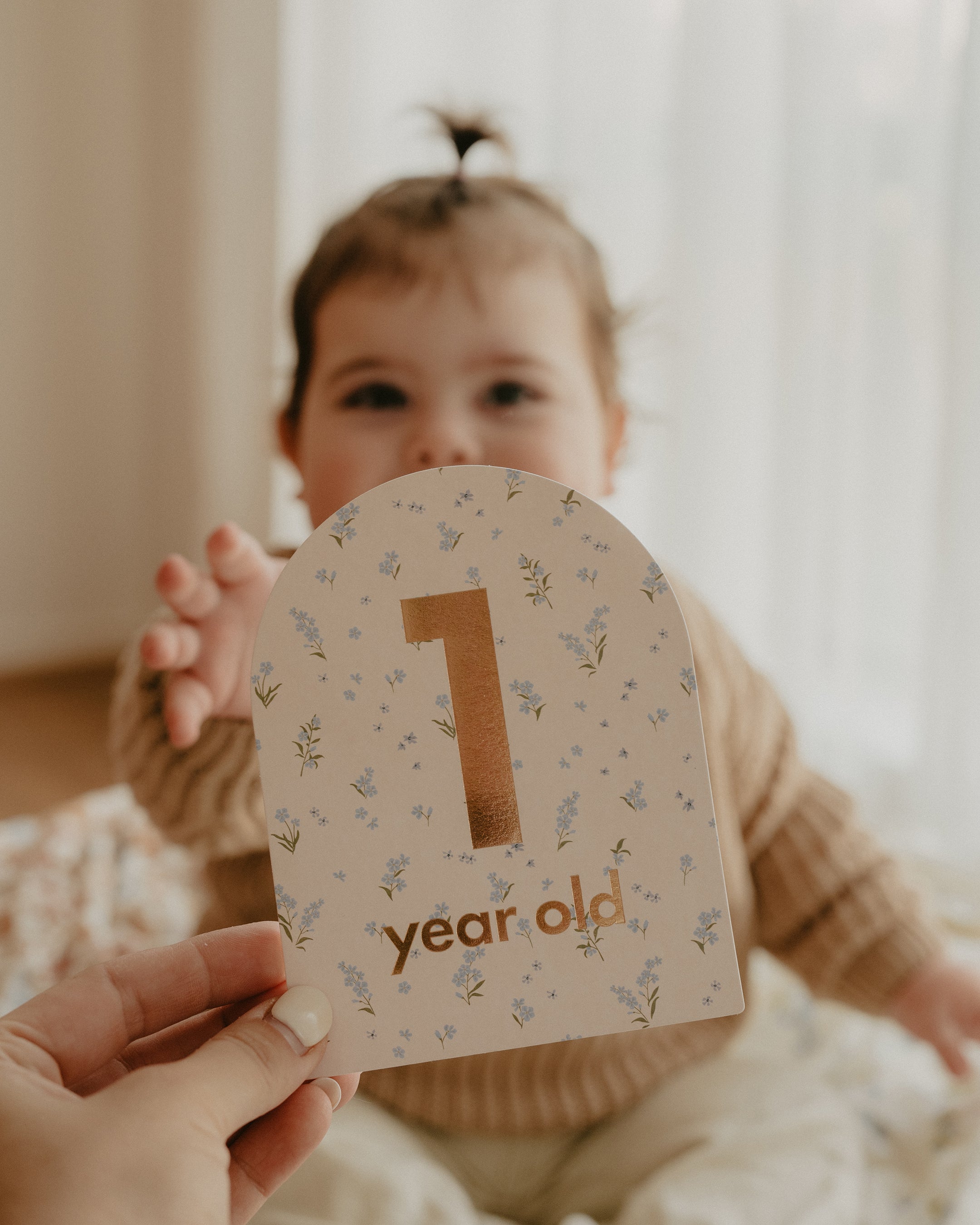 A baby with a small ponytail reaches towards the camera. In the foreground, a hand holds a charming FOX & FALLOW Baby Milestone Card Broderie that reads "1 year old" adorned with a floral design. The background is softly lit with natural light coming through sheer curtains, capturing this important milestone beautifully.