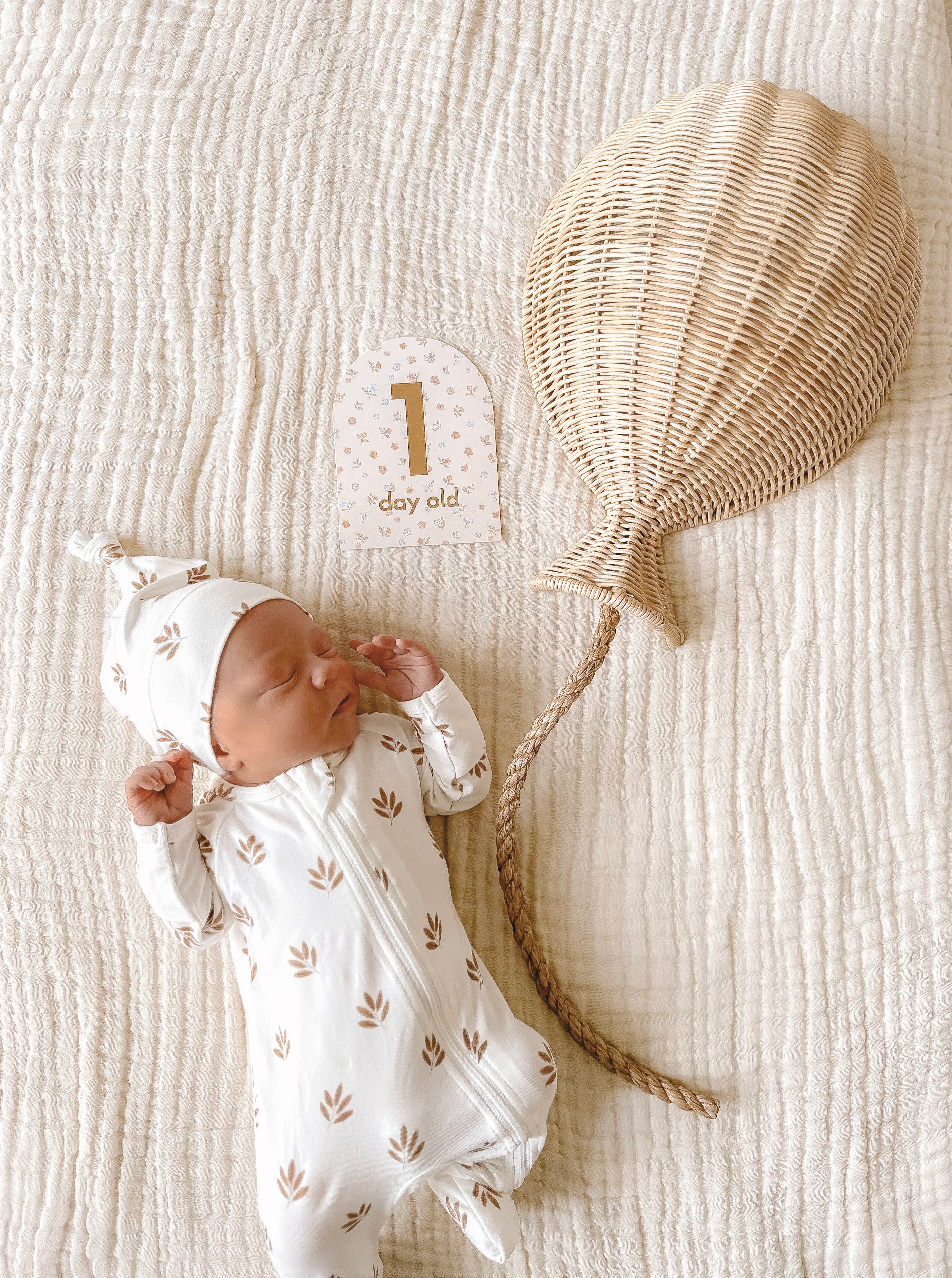 A sleeping newborn dressed in a white onesie with small brown leaf patterns and a matching hat lies on a white quilted blanket. Next to the baby is a FOX & FALLOW Baby Milestone Card Broderie that reads "1 day old" and a decorative, woven rattan balloon—perfect for capturing important milestones in your baby's birth announcement.