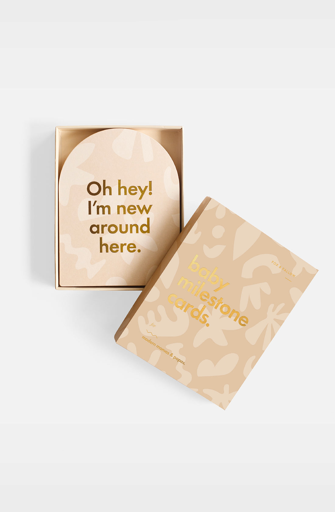 An open FOX & FALLOW Helios Baby Milestone Cards box in beige with gold "baby milestone cards" text displays a card saying "Oh hey! I'm new around here." The packaging has abstract light brown and cream patterns.