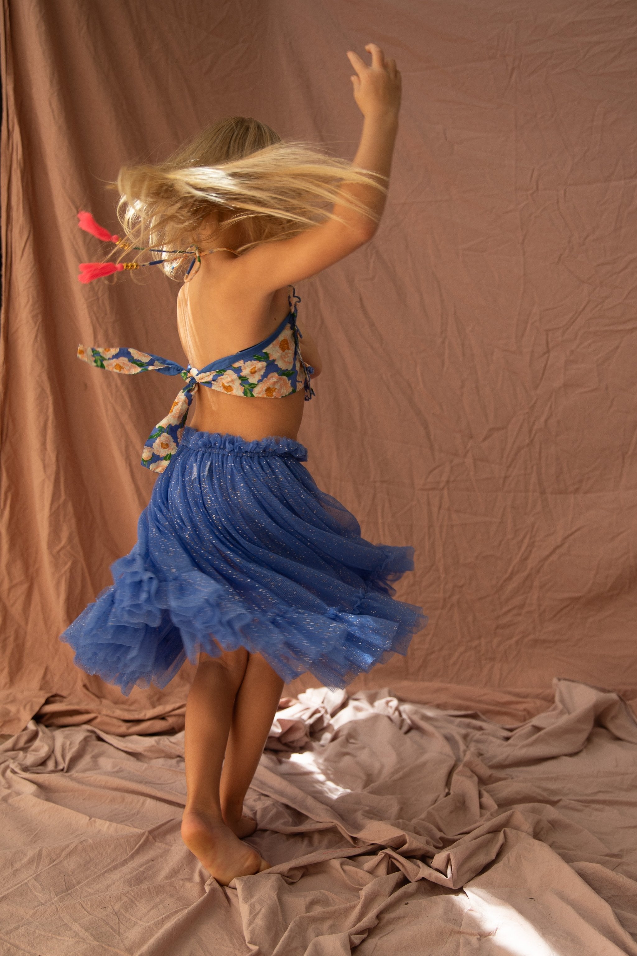 A child wearing a floral top and the Carrie Tutu in Bondi Blue by BELLA + LACE is twirling with arms raised, creating a dynamic motion effect. The background is composed of gently crumpled neutral-toned fabric, enhancing the whimsical feel of the scene.