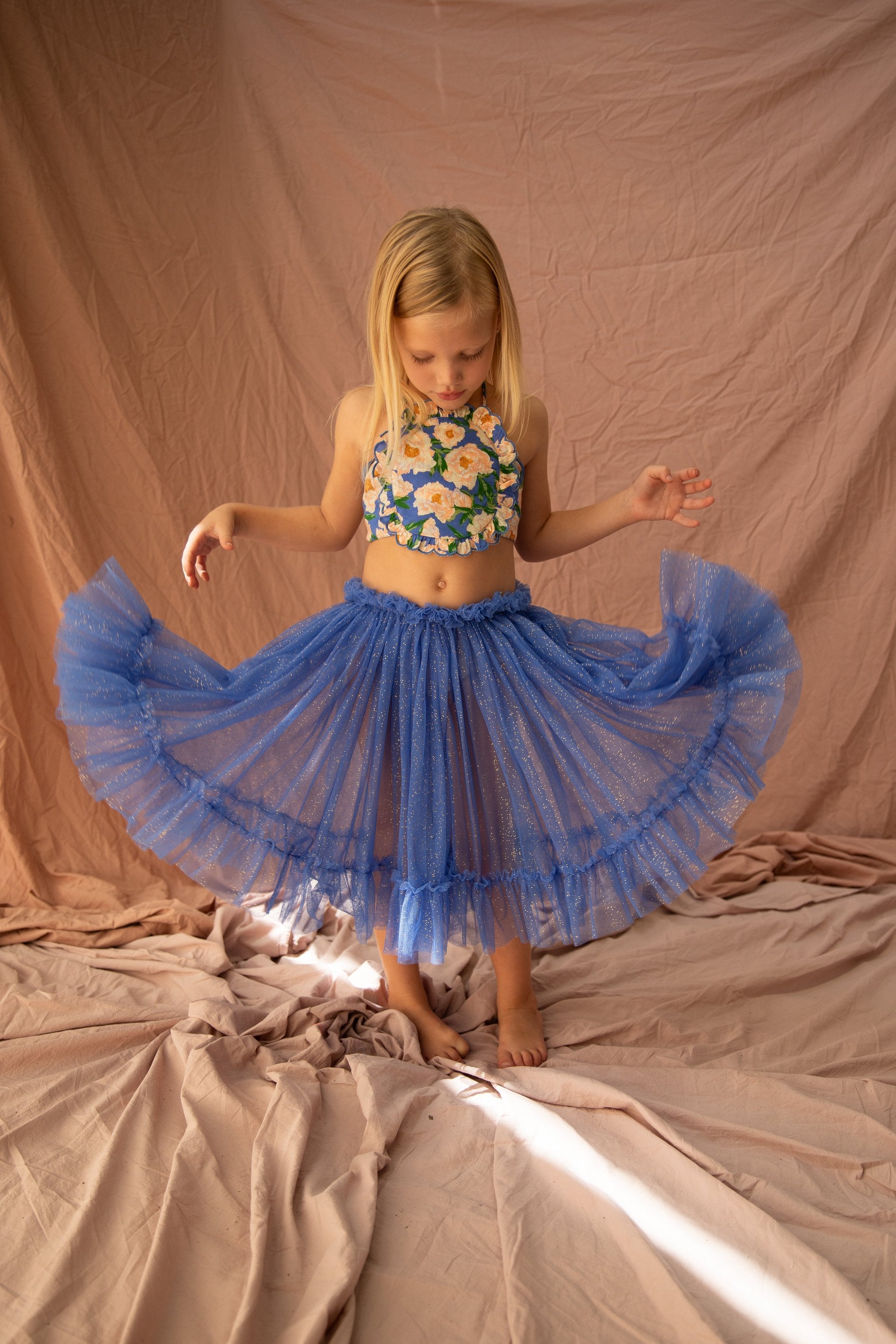 A young girl stands on a crinkled fabric backdrop, wearing a flower-patterned halter top and a sparkly, blue tulle skirt. With the grace of a BELLA + LACE Carrie Tutu Bondi Blue design, she holds the sides of her skirt, spreading it out and looking down. Sunlight filters in, casting soft shadows on the fabric behind her.