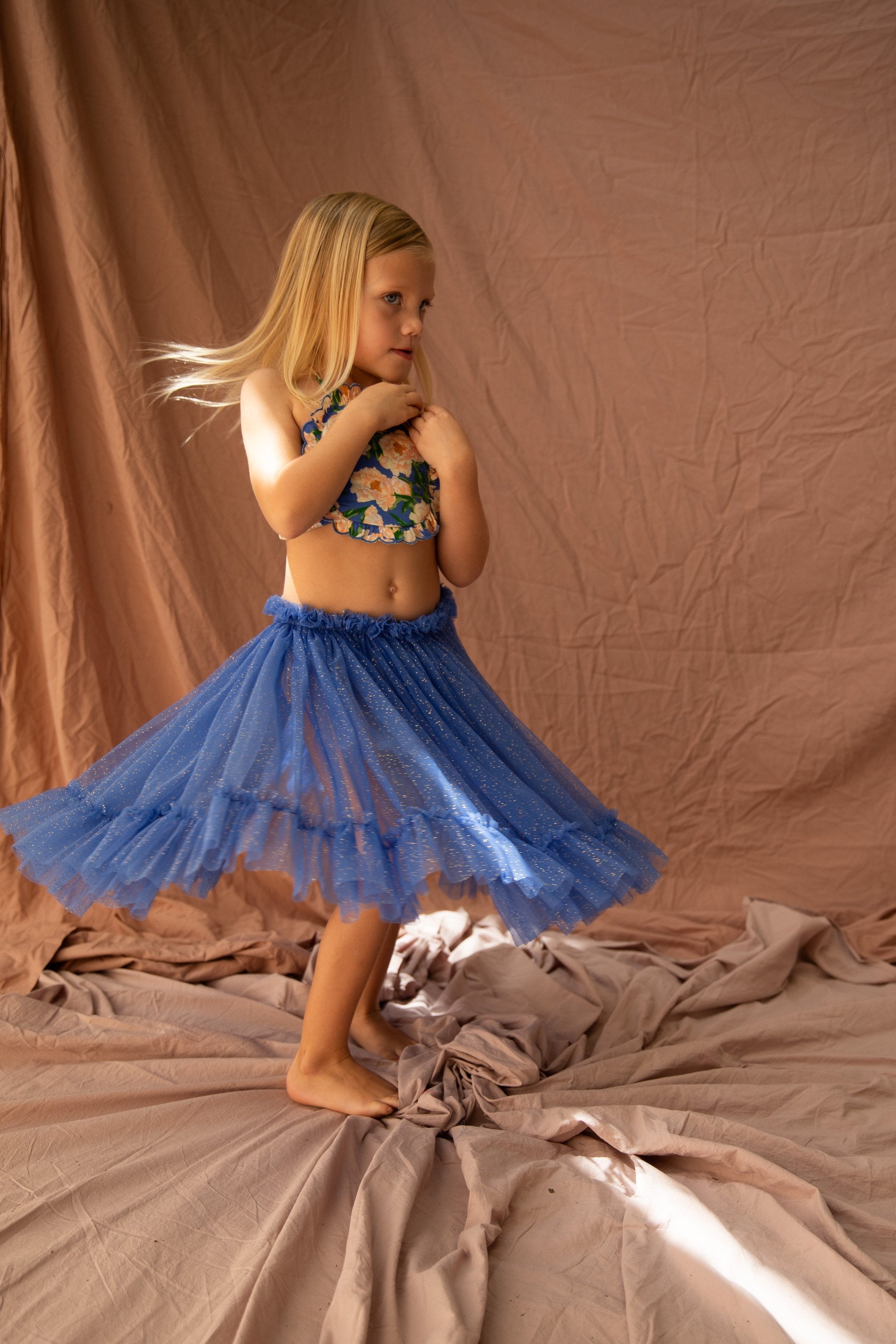 A young girl with long blonde hair is twirling in a sparkly Bondi Blue Carrie Tutu from BELLA + LACE in front of a beige backdrop. She is wearing a sleeveless floral top and is barefoot, with a playful and thoughtful expression on her face. The fabric on the floor adds texture to the scene.