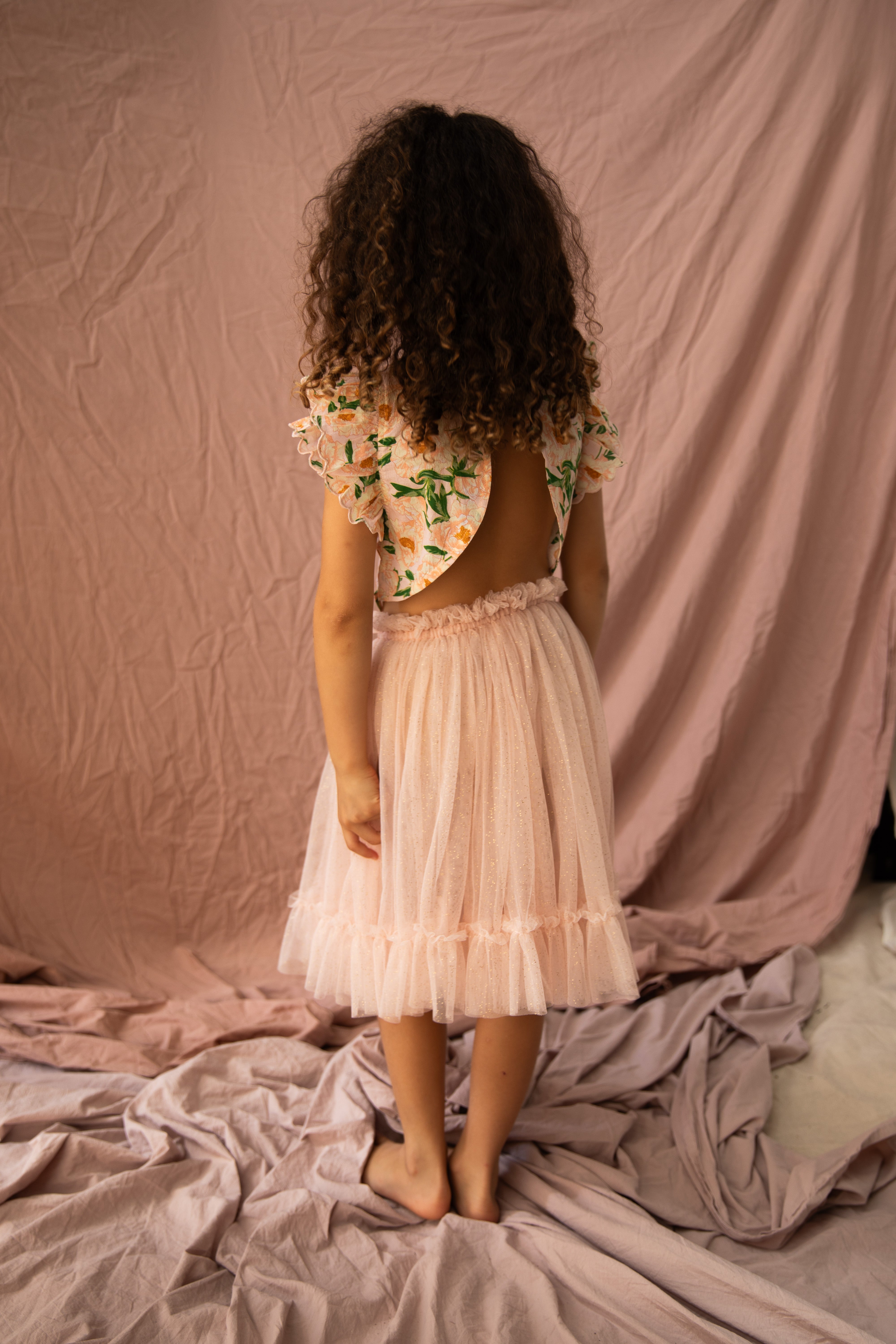 A young girl with curly hair stands facing away in a BELLA + LACE Carrie Tutu Strawberry Sundae dress, featuring a soft tulle skirt and an open-back top adorned with floral embroidery. The backdrop consists of gently draped pink fabrics.