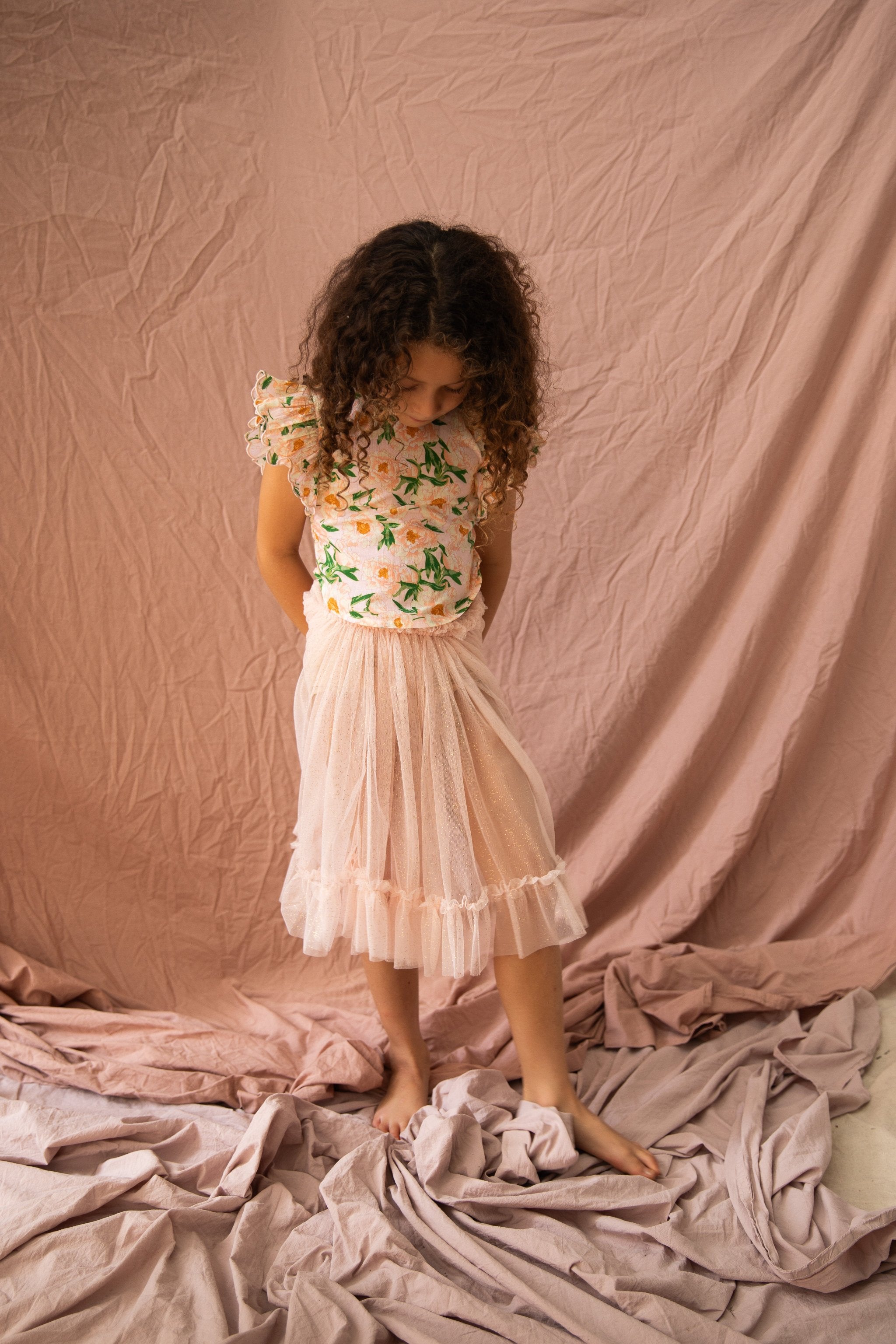 A young child with curly hair, wearing a BELLA + LACE floral top and a Carrie Tutu in Strawberry Sundae, stands barefoot on crumpled fabric. The backdrop is also a soft pink fabric. The child looks down with hands clasped behind their back.