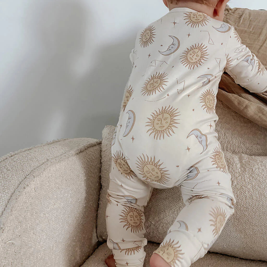 A baby wearing the "Celestial Zip Jumpsuit Cream" from BAM LOVES BOO, showcasing celestial sun, moon, and star patterns on cream-colored fabric, is playfully climbing on a beige couch. The outfit is crafted from bamboo organic cotton and reveals only part of the back and legs as the baby faces away.