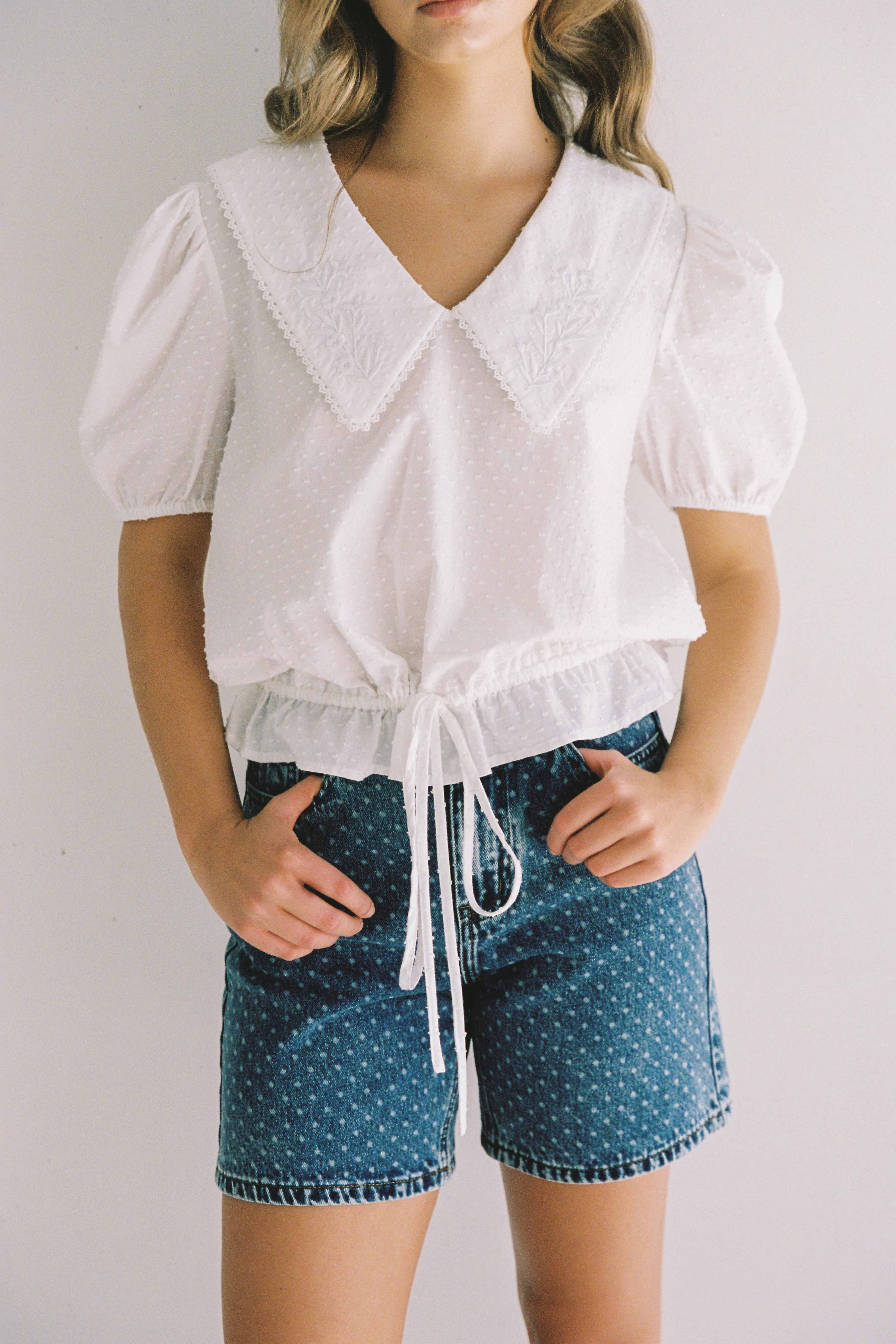 A person wearing a white blouse with a wide, embroidered collar and puff sleeves paired with high-waisted Bud Denim Short Spot in blue cotton denim by JUNI stands against a plain white background. The blouse is cinched at the waist with a drawstring. The person's hands rest on the functional pockets of their shorts.