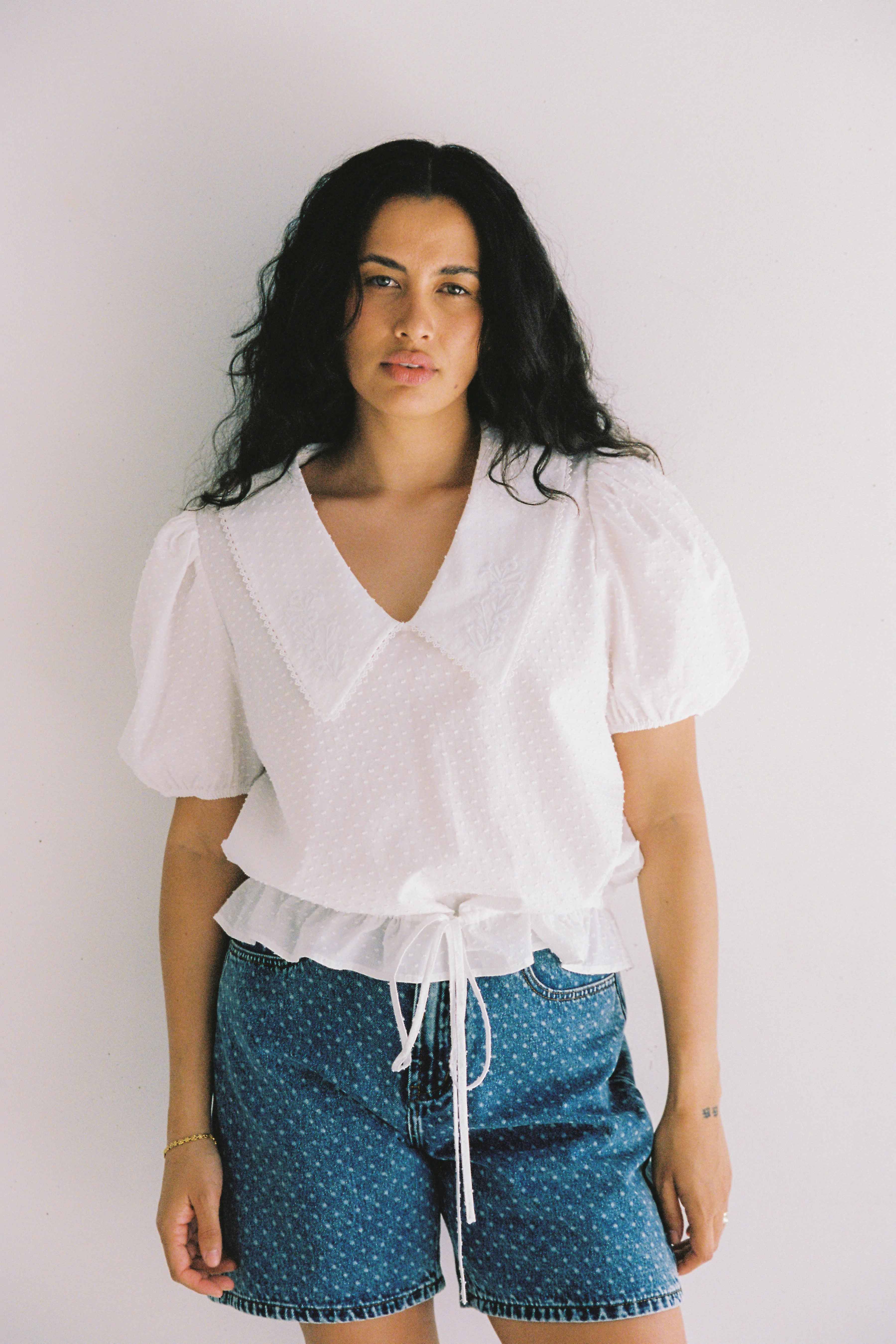 A woman with long, wavy hair wearing a white blouse featuring puffed short sleeves and a V-neck collar is paired with the JUNI Bud Denim Short Spot high-waist blue denim shorts. She stands against a plain white background, looking directly at the camera.