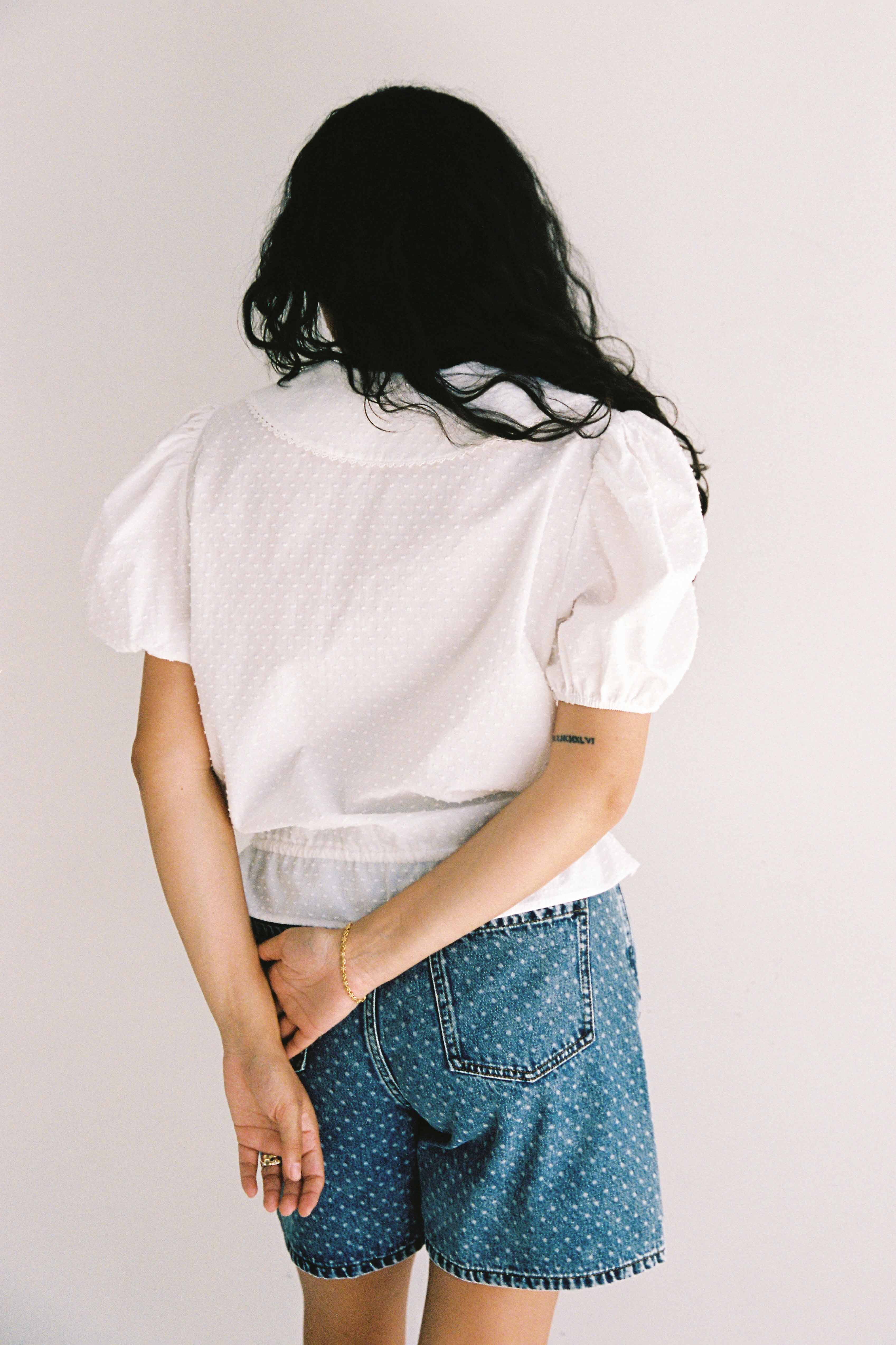 A person with long, dark hair stands with their back to the camera, wearing a white short-sleeved blouse and JUNI's Bud Denim Short Spot, featuring functional pockets. With one arm hanging down and the other bent behind their back, they showcase rings and a bracelet against a plain wall backdrop.