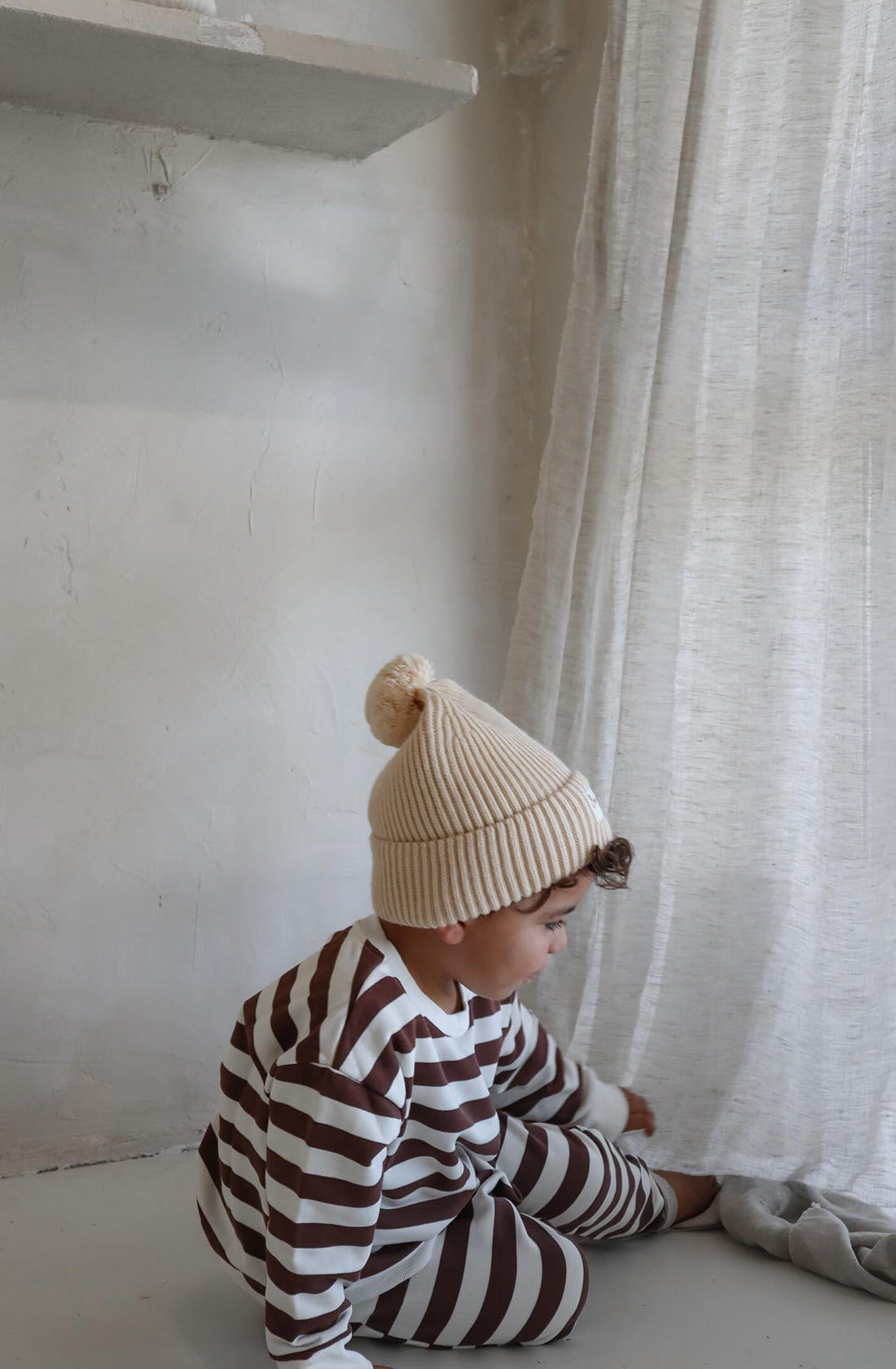 A young toddler wearing the Camden chocolate brown striped pullover and matching pants with the Bobby G cream beanie playing with window drapes.