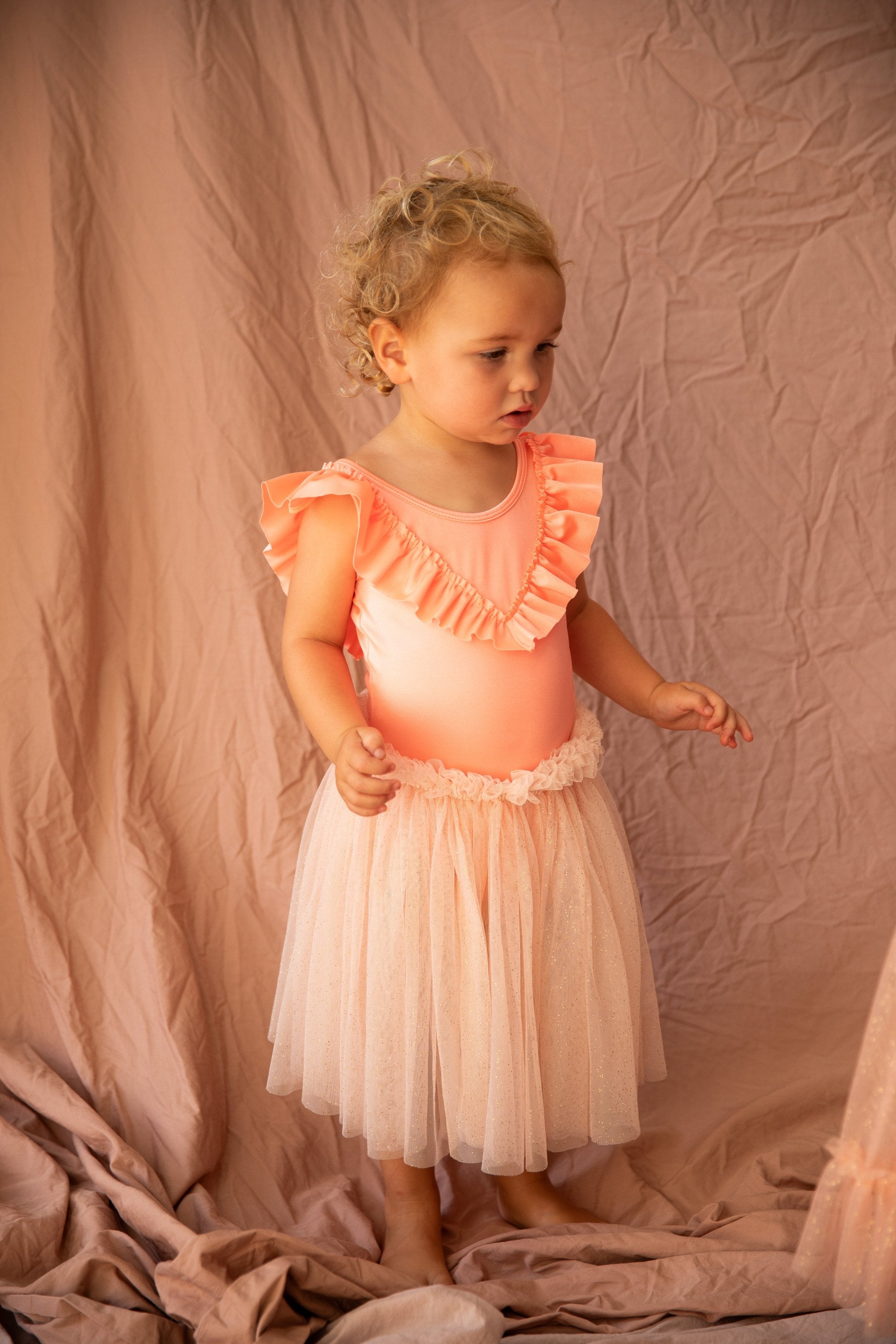 A toddler with curly blonde hair is in a BELLA + LACE Classic Tutu Strawberry Sundae - Size 1-2, featuring ruffled sleeves and a tulle skirt. The child stands on a fabric backdrop, gazing thoughtfully to the right, evoking an enchanting BELLA + LACE moment.