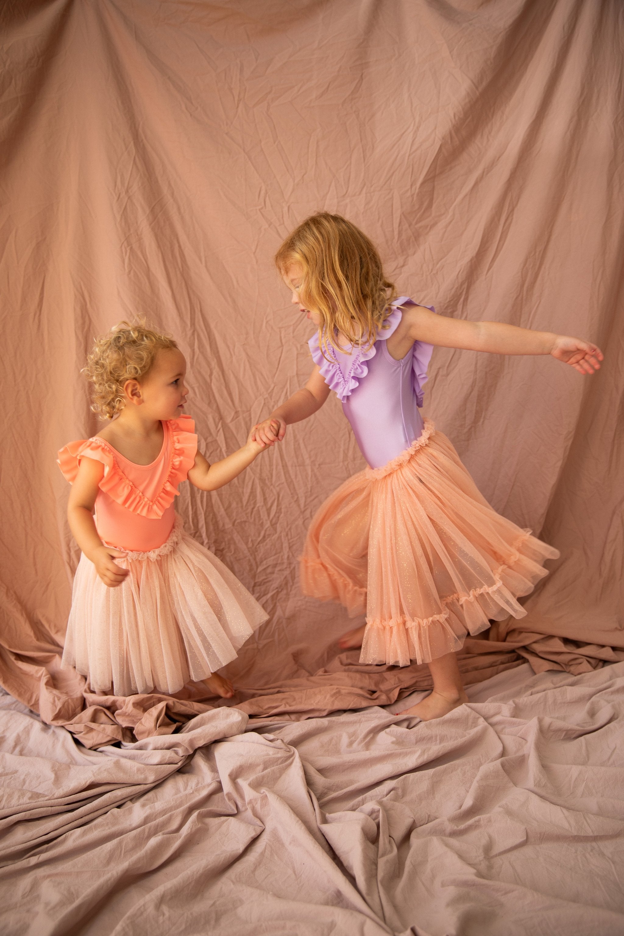 Two young children dressed in colorful outfits, one in a Classic Tutu Strawberry Sundae from BELLA + LACE and the other in a purple top with an extra soft tulle skirt. They hold hands and appear to be dancing on a fabric-covered surface with a draped fabric background, embodying the charm of BELLA + LACE.