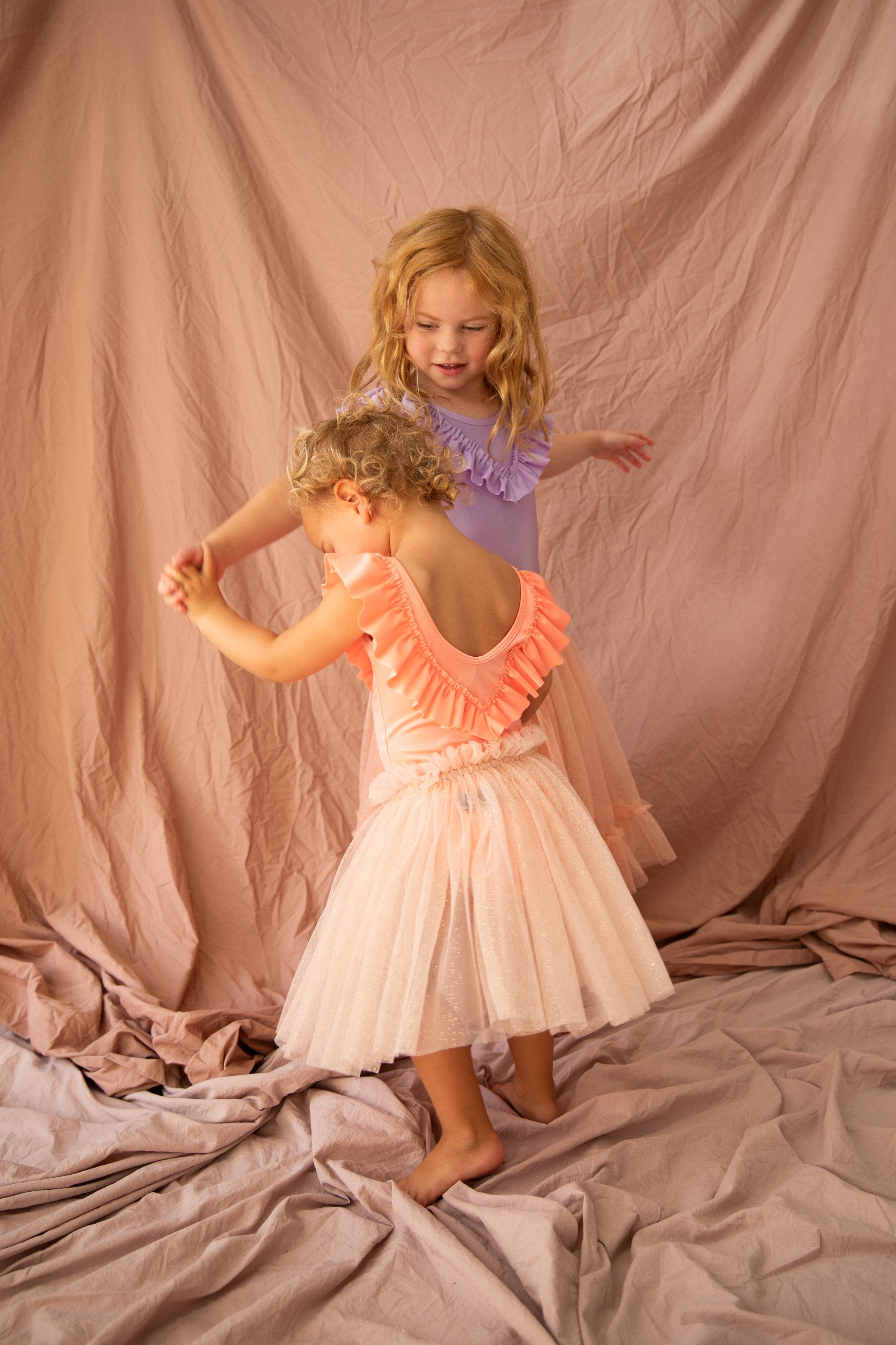 Two young children dance on a soft, crumpled fabric backdrop. One wears a peach ruffled dress, and the other wears the BELLA + LACE Classic Tutu Strawberry Sundae, Size 1-2. They hold hands and smile amidst gentle pink hues.