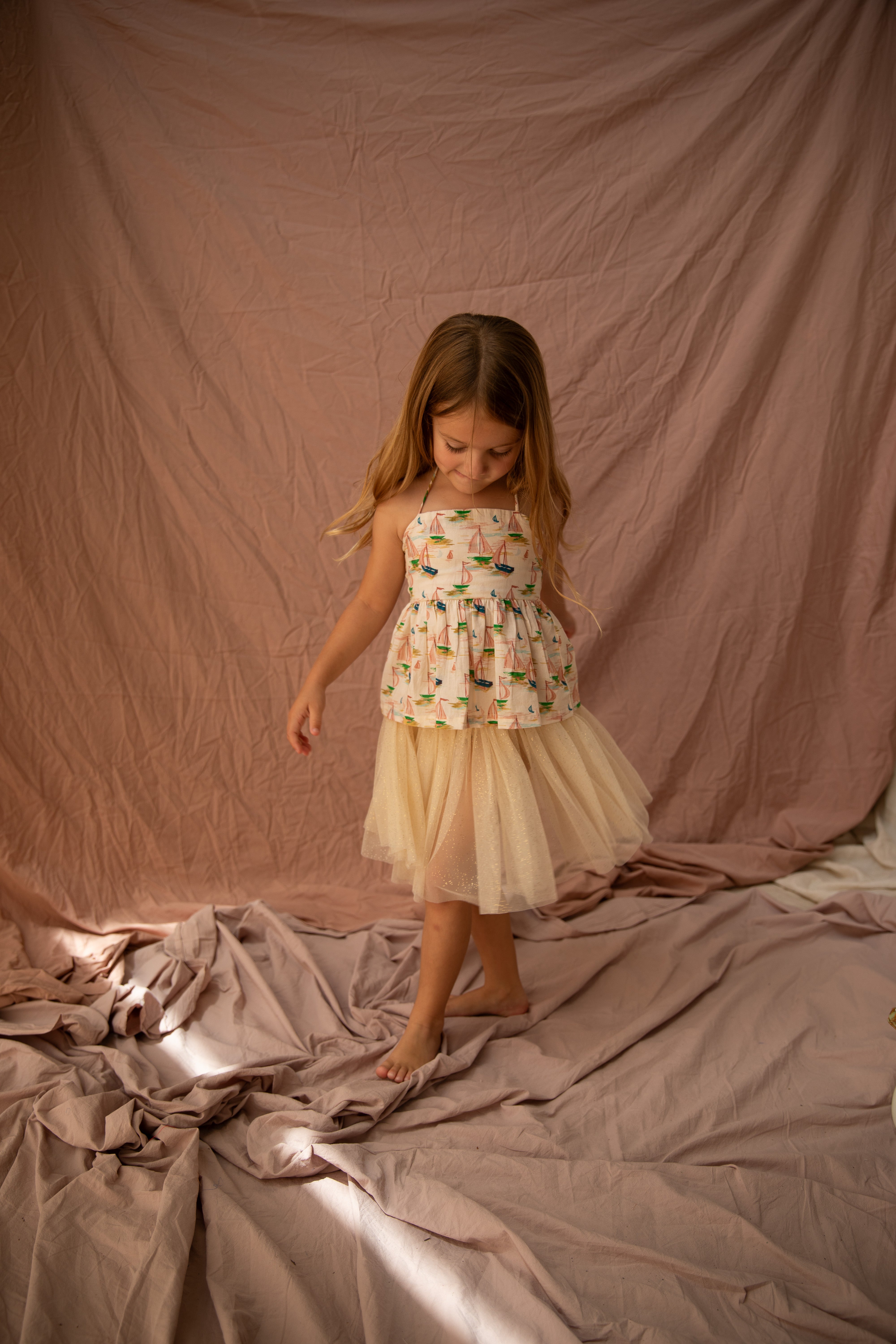 A young girl with long hair wears the BELLA + LACE's Classic Tutu Vanilla, a sleeveless, layered cream dress adorned with a floral pattern. She stands on textured, draped fabric with a thoughtful expression, set against a soft beige backdrop.