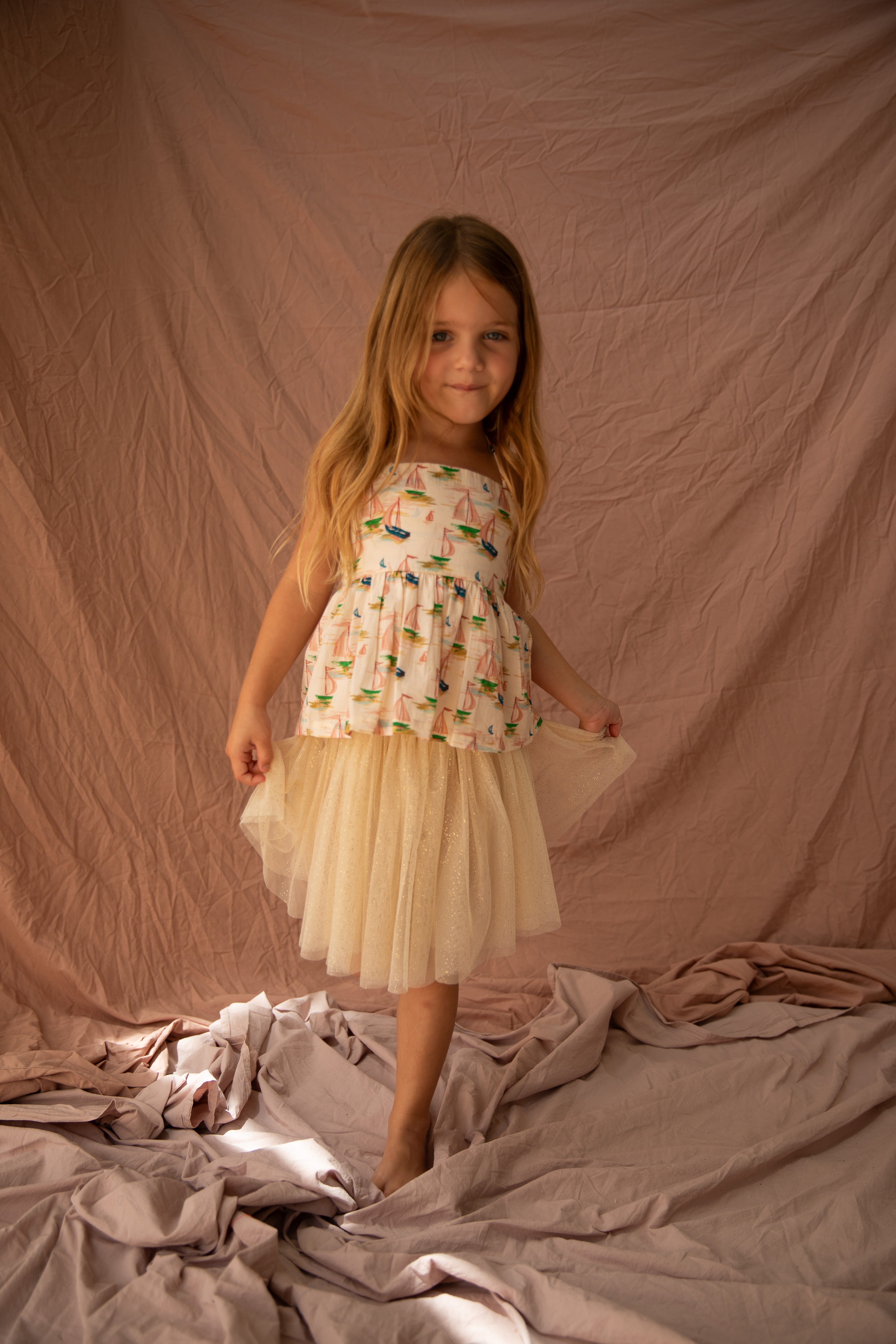 A young girl with long hair stands gracefully on crumpled fabric, wearing the Classic Tutu Vanilla by BELLA + LACE, which features a colorful pattern. The beige backdrop and soft lighting create a warm, gentle atmosphere.