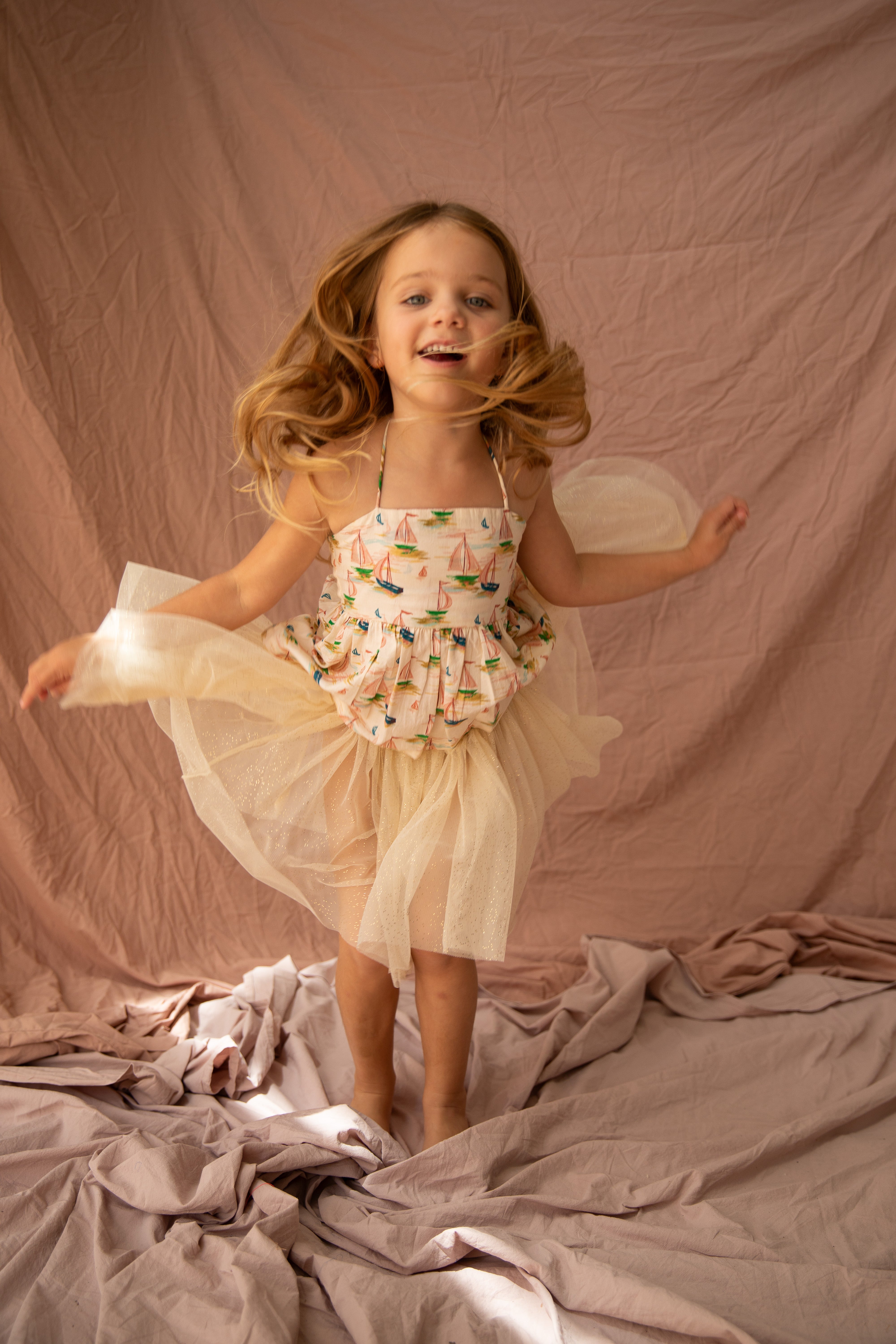 A young girl with long hair is joyfully jumping on a soft, wrinkled fabric surface. She is wearing the Classic Tutu Vanilla from BELLA + LACE, adorned with sailboat prints and delicate lace accents. The background is a muted pink cloth, and her expression is cheerful and energetic.