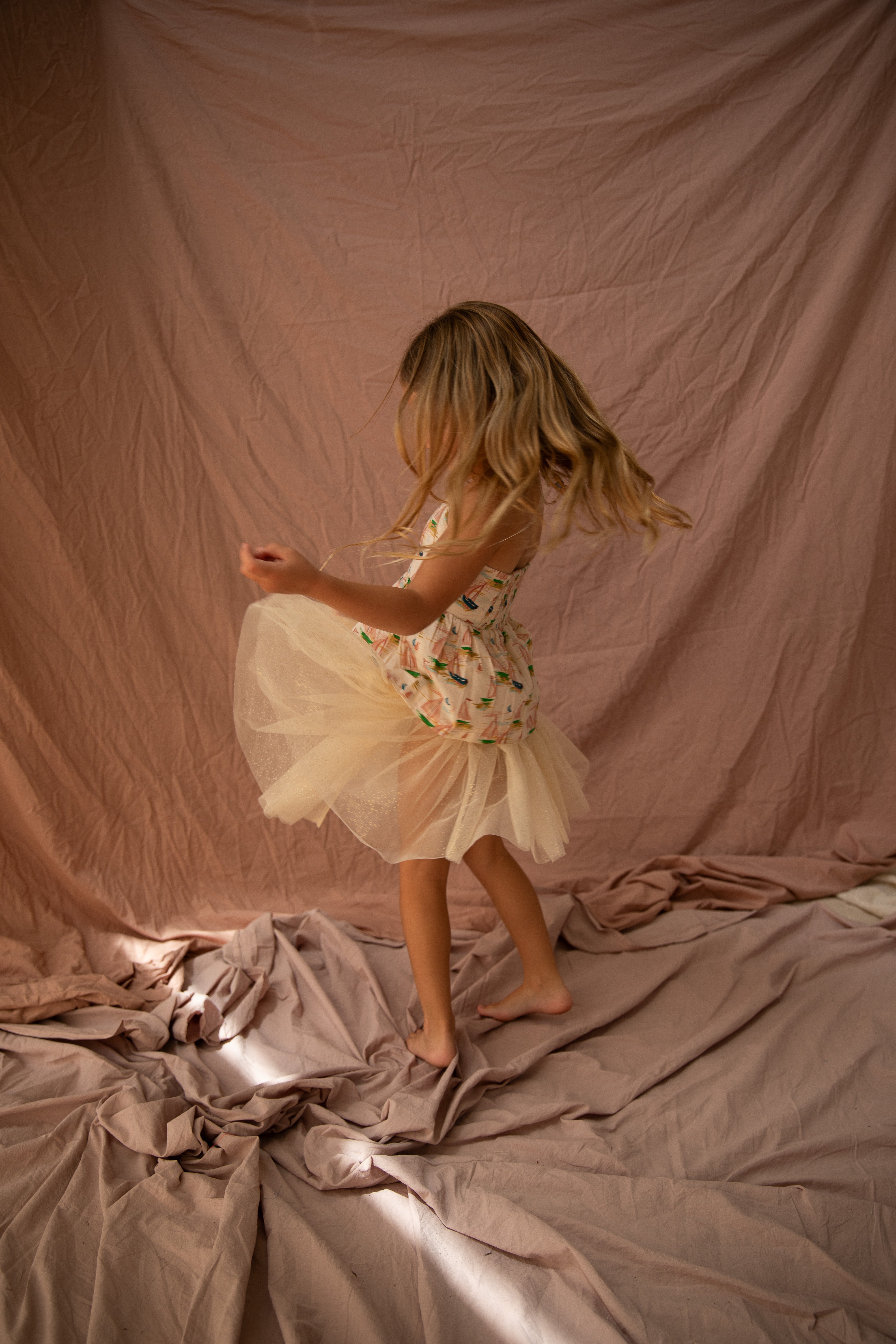 A young girl with long blonde hair twirls joyfully in a BELLA + LACE Classic Tutu Vanilla dress with a soft tulle skirt. She is barefoot, surrounded by crumpled fabric in a warmly lit room, creating a peaceful and whimsical atmosphere.