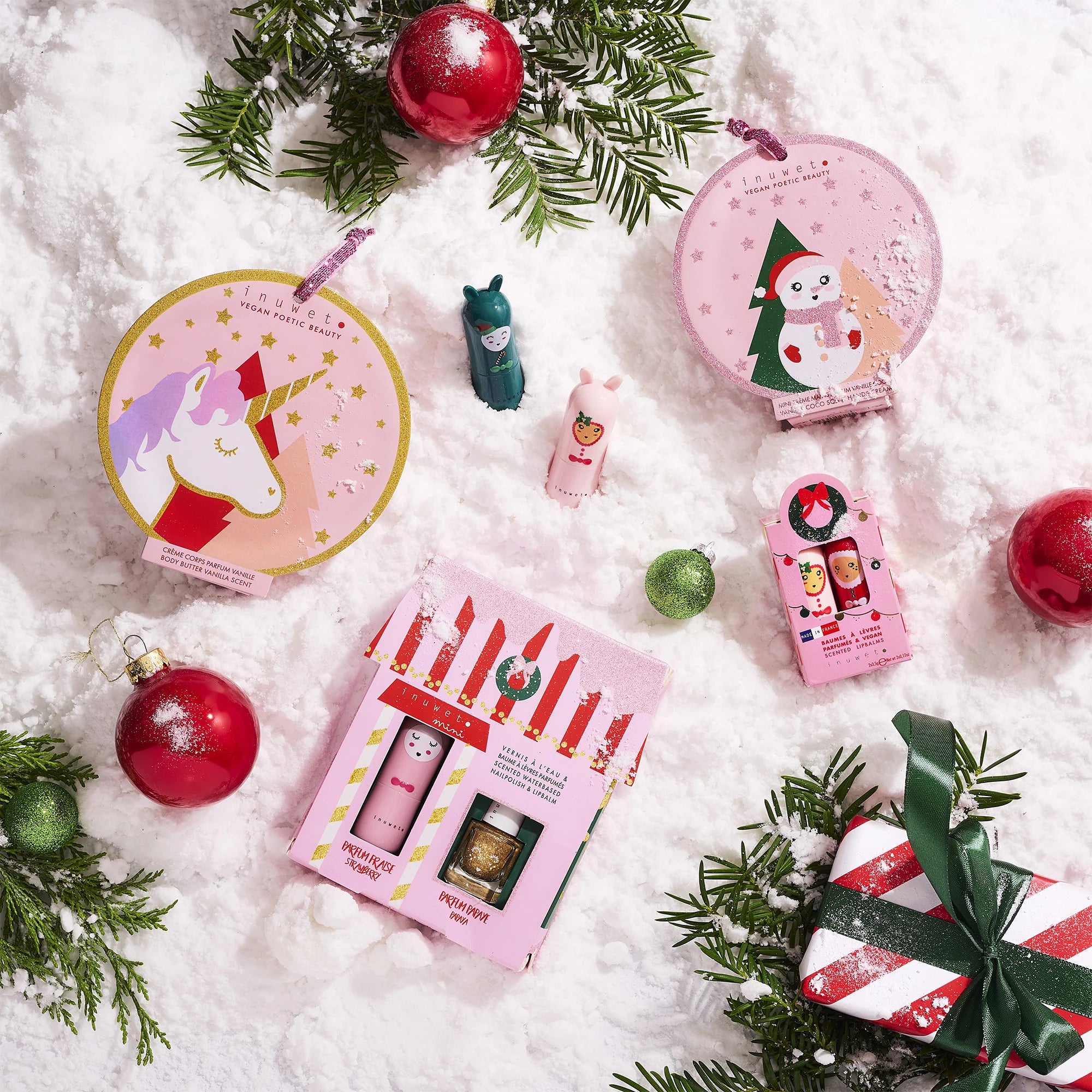 A charming display of festive beauty products arranged on snow with fir branches and red ornaments, featuring circular unicorn and snowman-themed tins, a small bottle, and the La Maison Juliette Gift Box by INUWET with playful designs. A wrapped gift nearby offers vegan formulas like strawberry-scented lip balm for added cheer.