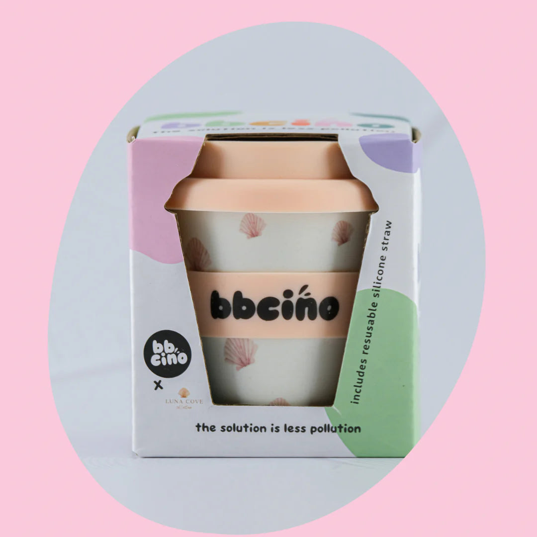 Image of a BBCINO Reusable Babycino Cup 120ml Cove (Limited Edition) in its packaging. The cup is white with pink accents and has a beige lid. The box is colorful with pastel shapes and text that says, "BBCINO" and "the solution is less pollution." It includes a reusable silicone straw made from bamboo fibers.