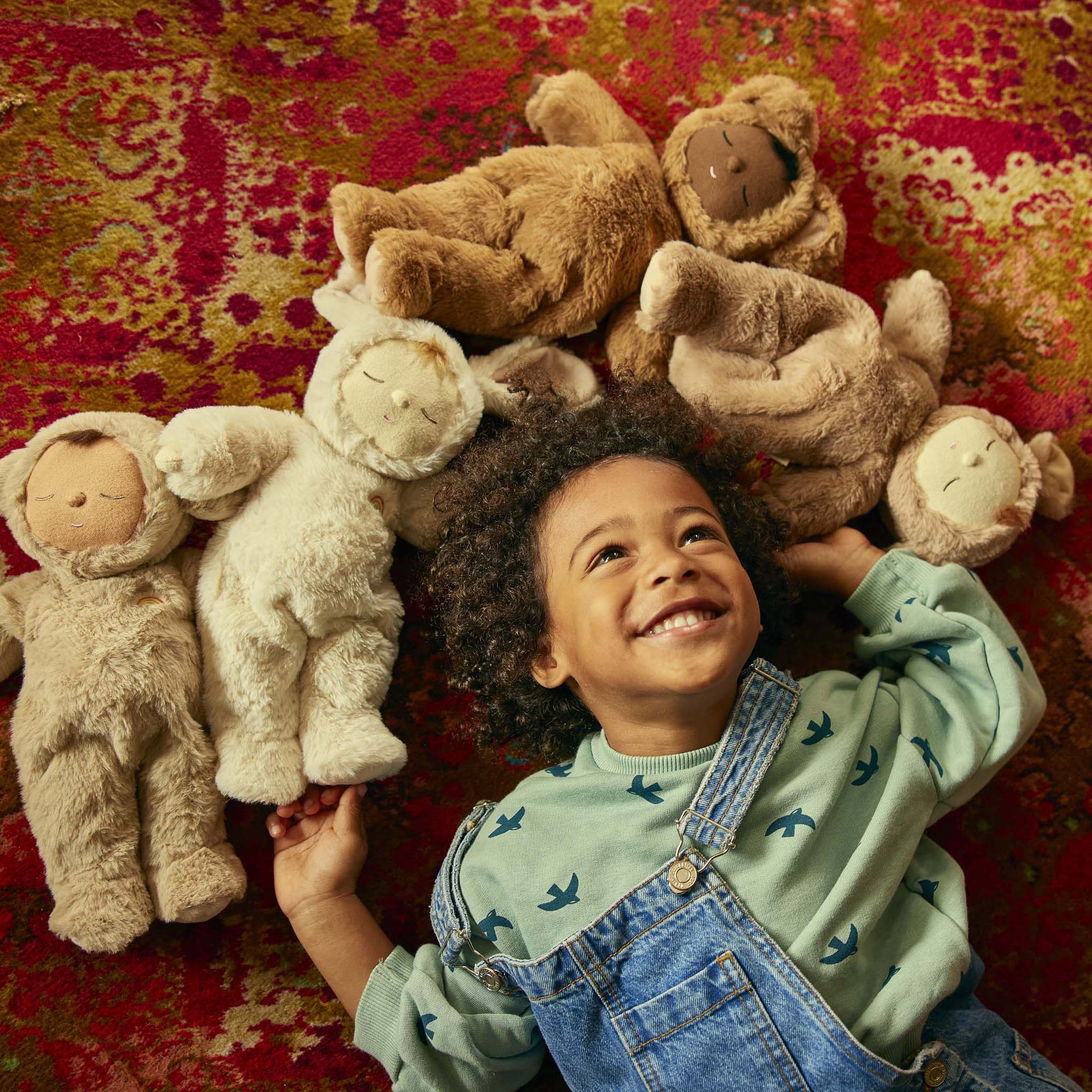 A smiling young child with curly hair lies on a colorful, patterned rug, cuddling a Cozy Dinkum Lamby Pip from OLLI ELLA amidst several plush teddy bears. The child is wearing a green sweater with bird prints and denim overalls, basking in the playful and cozy atmosphere created by the soft toys.