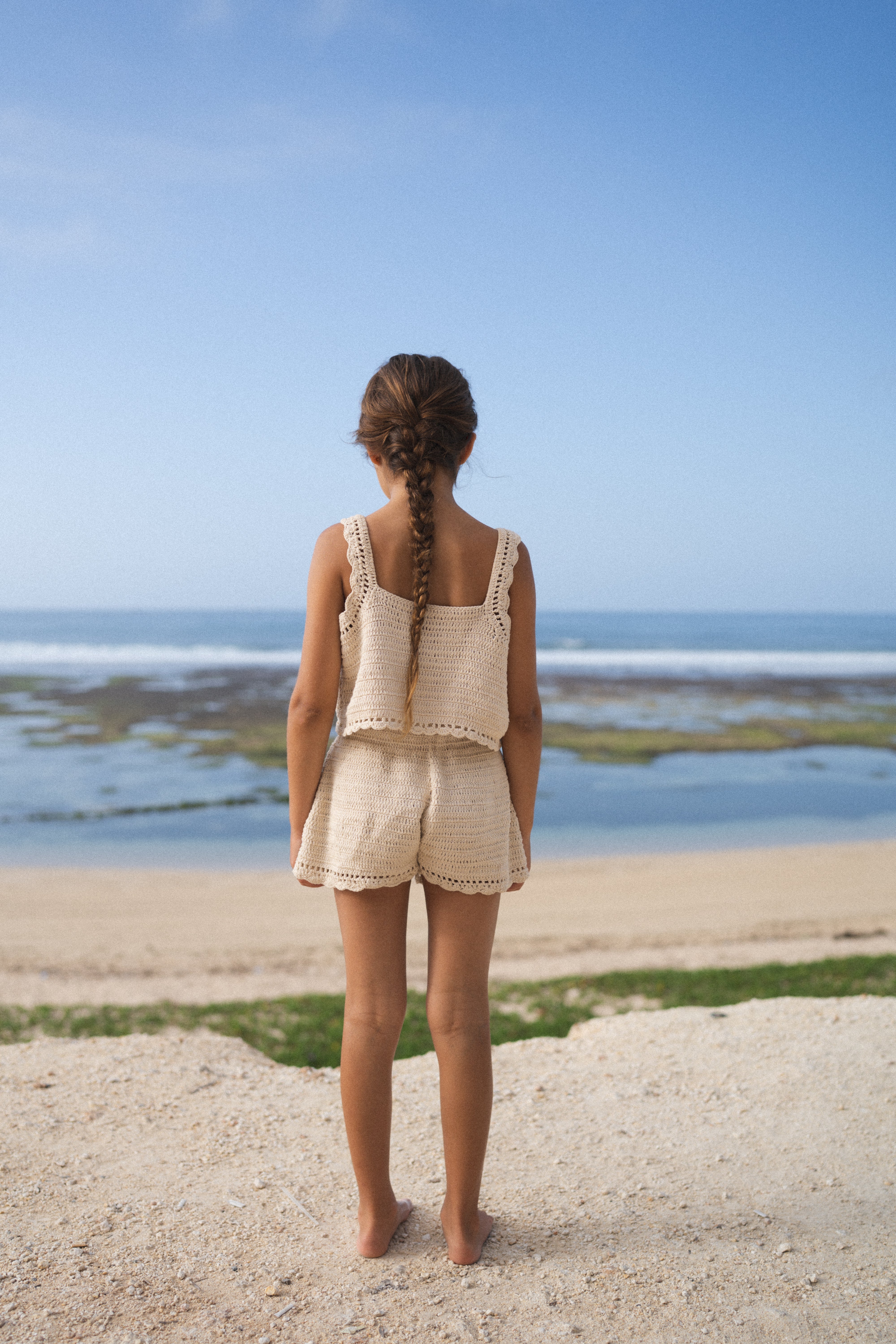 A young girl with long braided hair stands barefoot on a sandy beach, facing the ocean. She is wearing the Crochet Top Ecru by ILLOURA THE LABEL, a beige crochet top in a soft ecru colourway, paired with shorts. The sky is clear, and the sea appears calm with gentle waves.