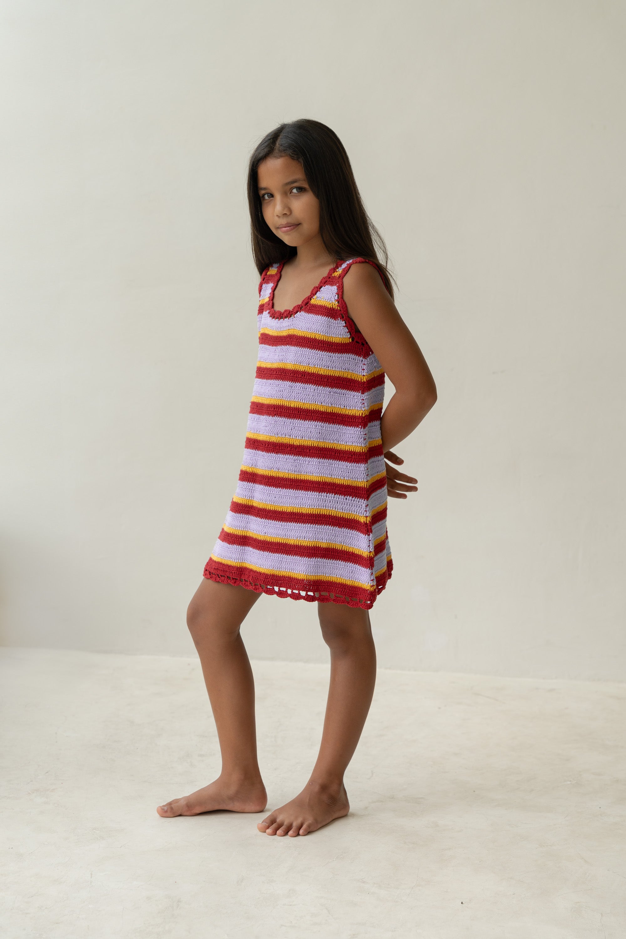 A young girl with long dark hair is wearing the Crochet Mini Sunset Stripe dress by Illoura The Label. She is barefoot on a light-colored floor against a plain background, arms positioned behind her back, gazing at the camera.