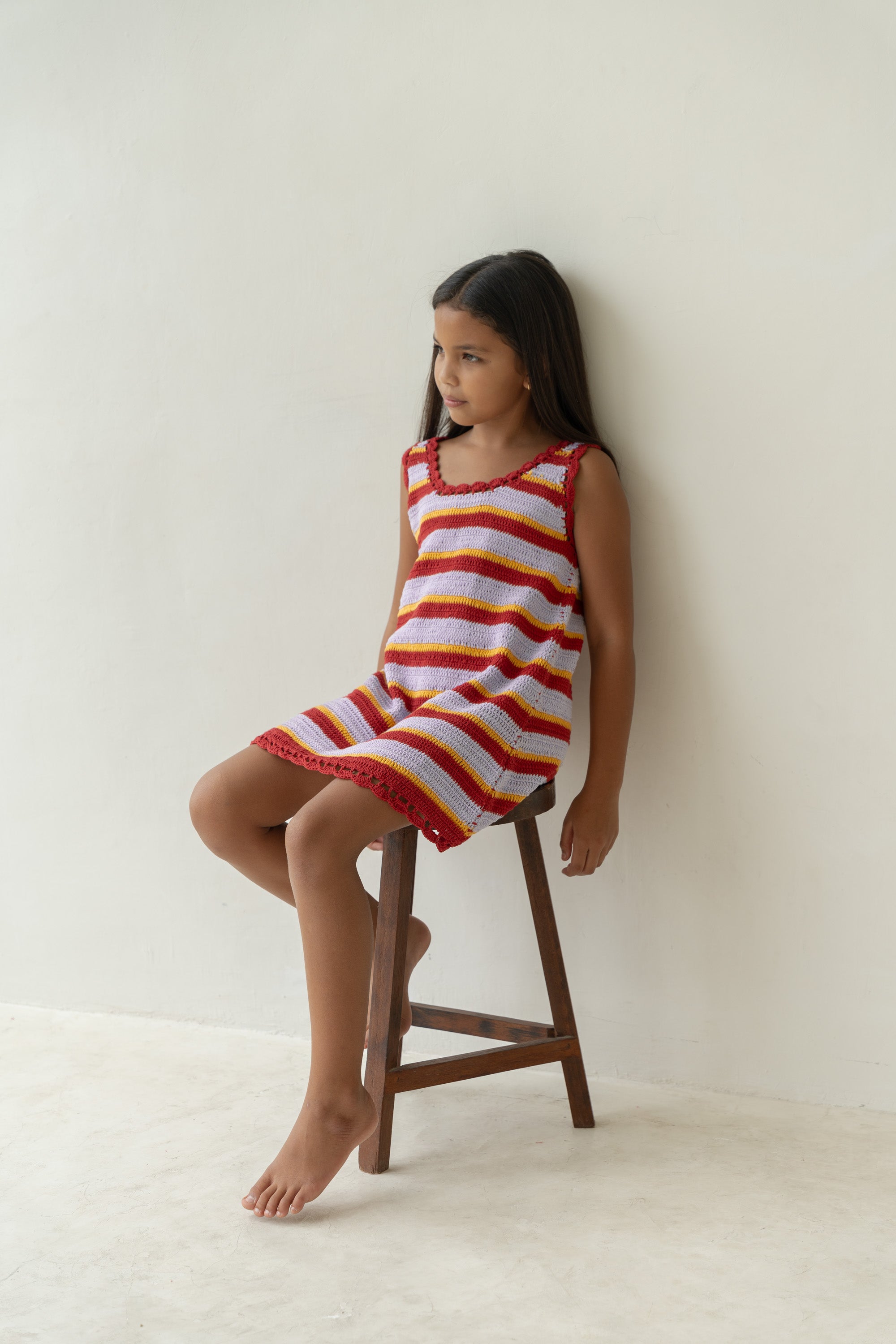 A young girl with long dark hair sits on a wooden stool against a plain white wall, wearing the Crochet Mini Sunset Stripe dress by ILLOURA THE LABEL. The vibrant crochet dress features horizontal bands in red, white, and yellow. Her expression is calm and relaxed.