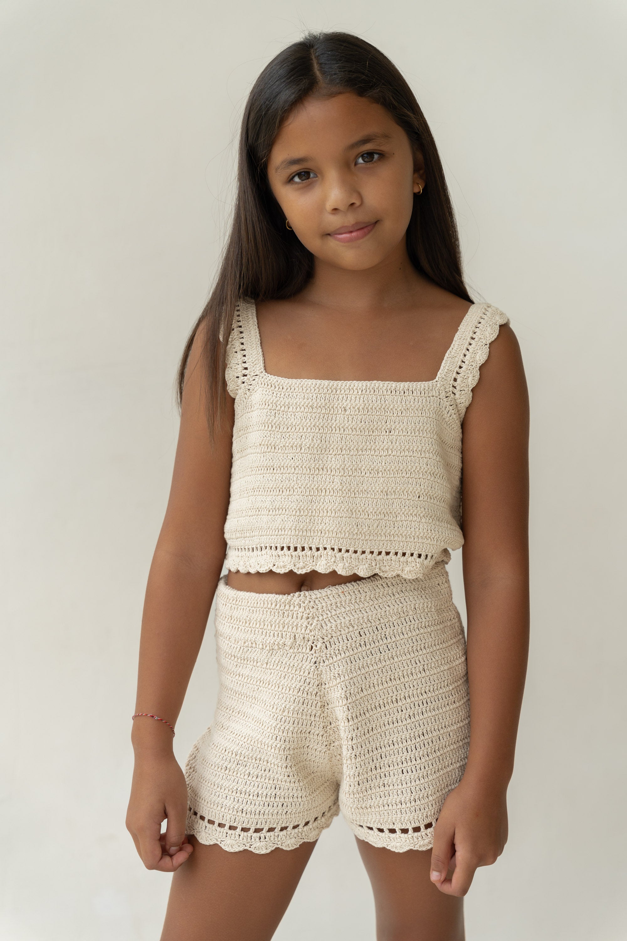 A young girl with long brown hair stands against a plain background, wearing an ILLOURA THE LABEL Crochet Top Ecru cotton tank top paired with matching shorts. Her hands rest by her sides as she maintains a neutral expression, embodying a simple yet elegant style.